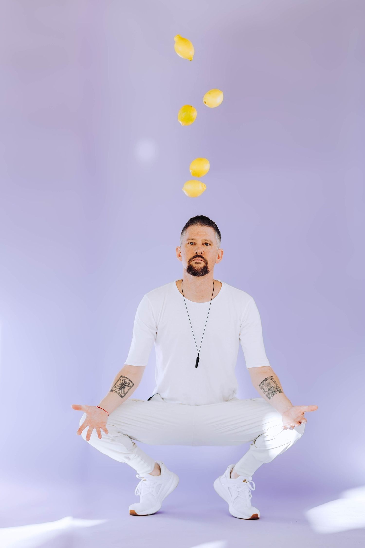 Tattooed man in white outfit squatting with lemons floating above his hands on purple background.