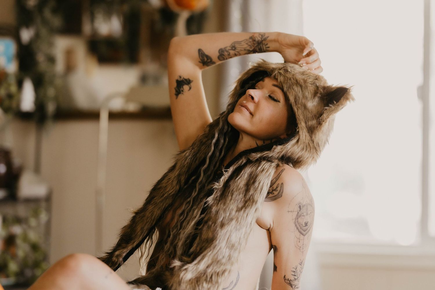Tattooed woman in a fur wolf hat sitting on a fur rug in a softly lit bohemian room.
