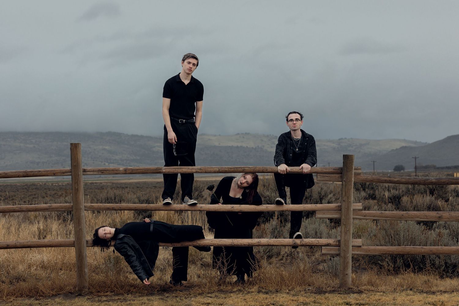Three people in black clothing pose dynamically around a wooden fence in a vast open field under stormy skies.