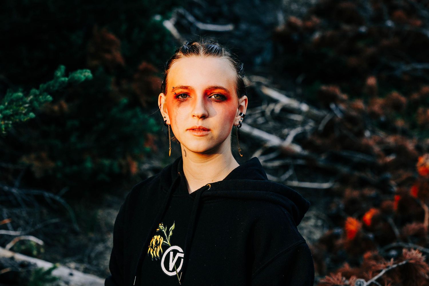 Young woman with bold red and orange eye makeup poses in black hoodie outdoors.