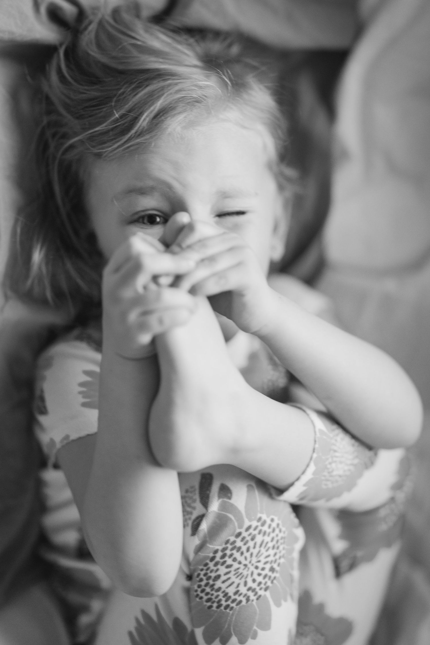 Black and white photo of a laughing toddler girl covering her mouth with her feet while lying down.