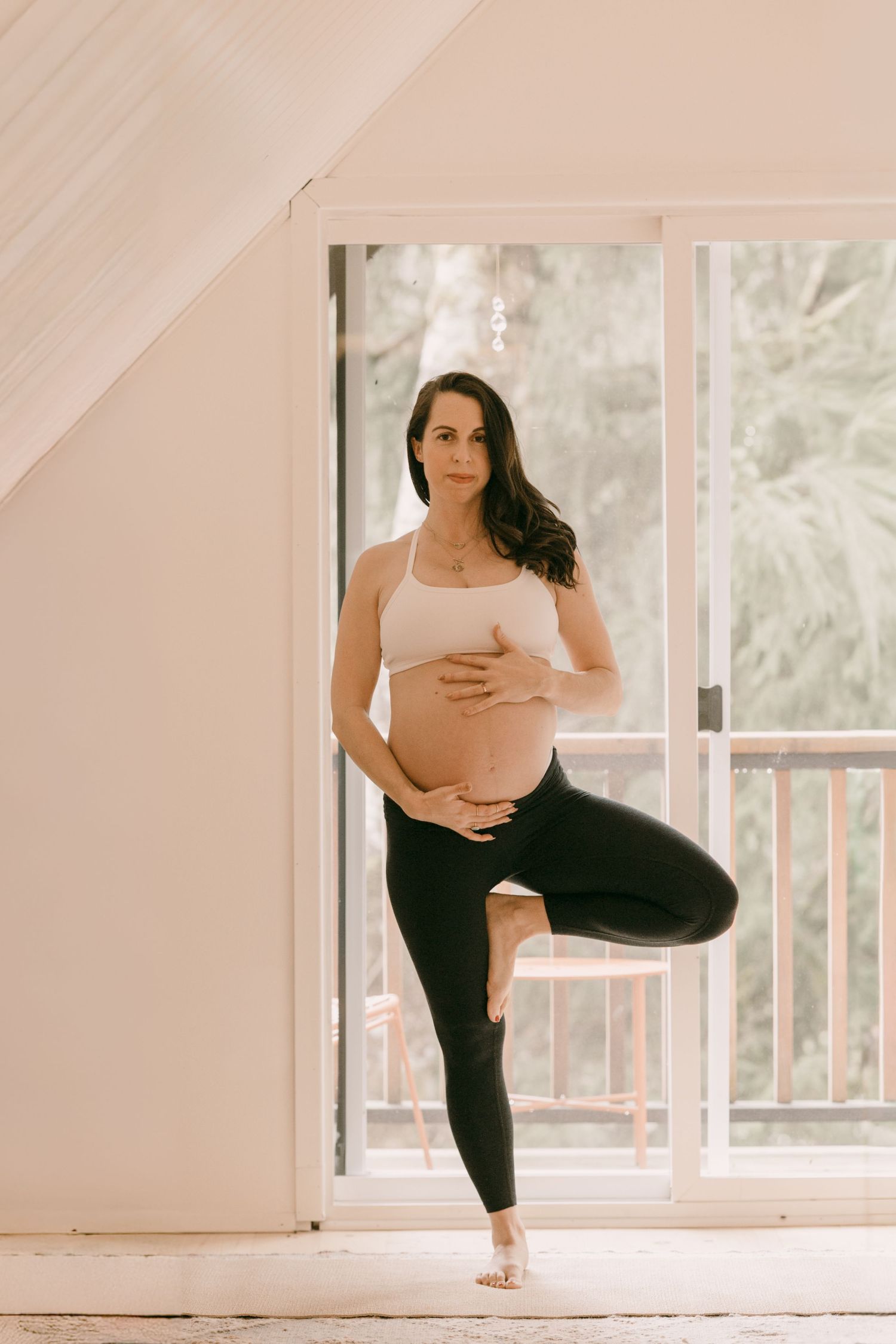 Pregnant woman in beige top and black leggings doing tree yoga pose indoors by a bright window.