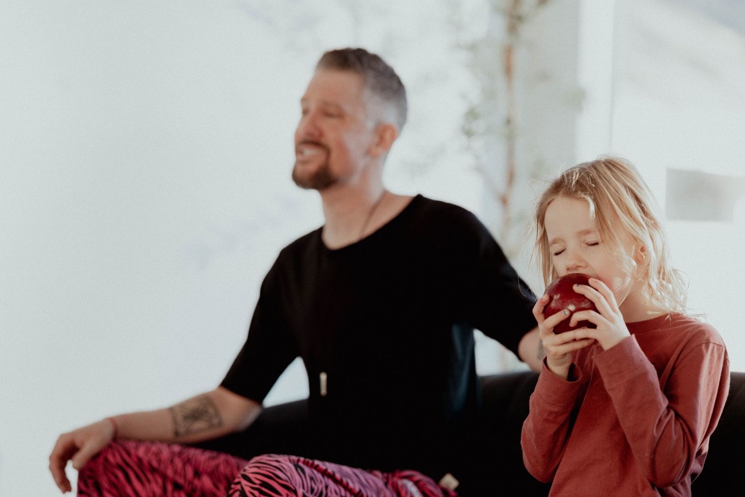 Tattooed man in black shirt meditating beside a young child eating a red apple indoors.