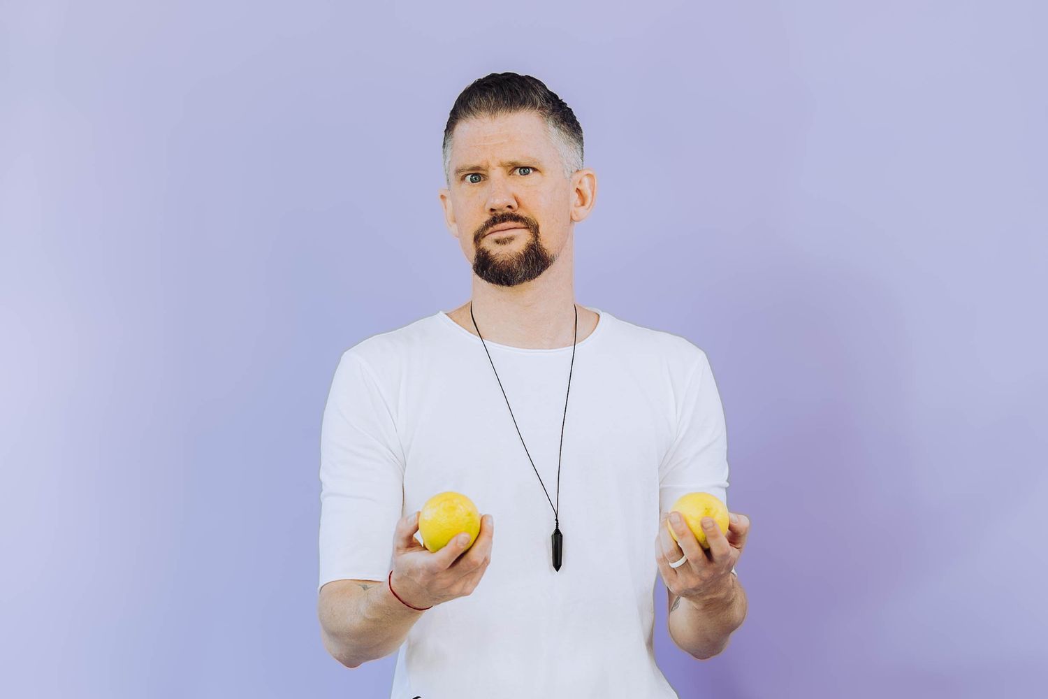 Bearded man in white t-shirt holding two yellow lemons against a light purple background.