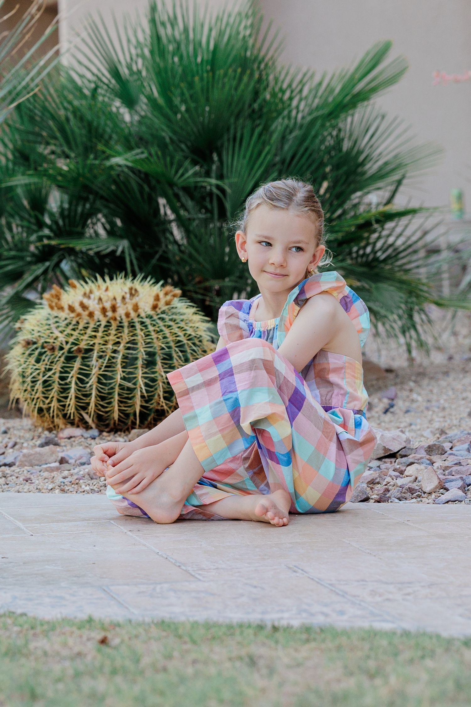 Young girl in colorful plaid dress sitting barefoot on patio near desert plants.