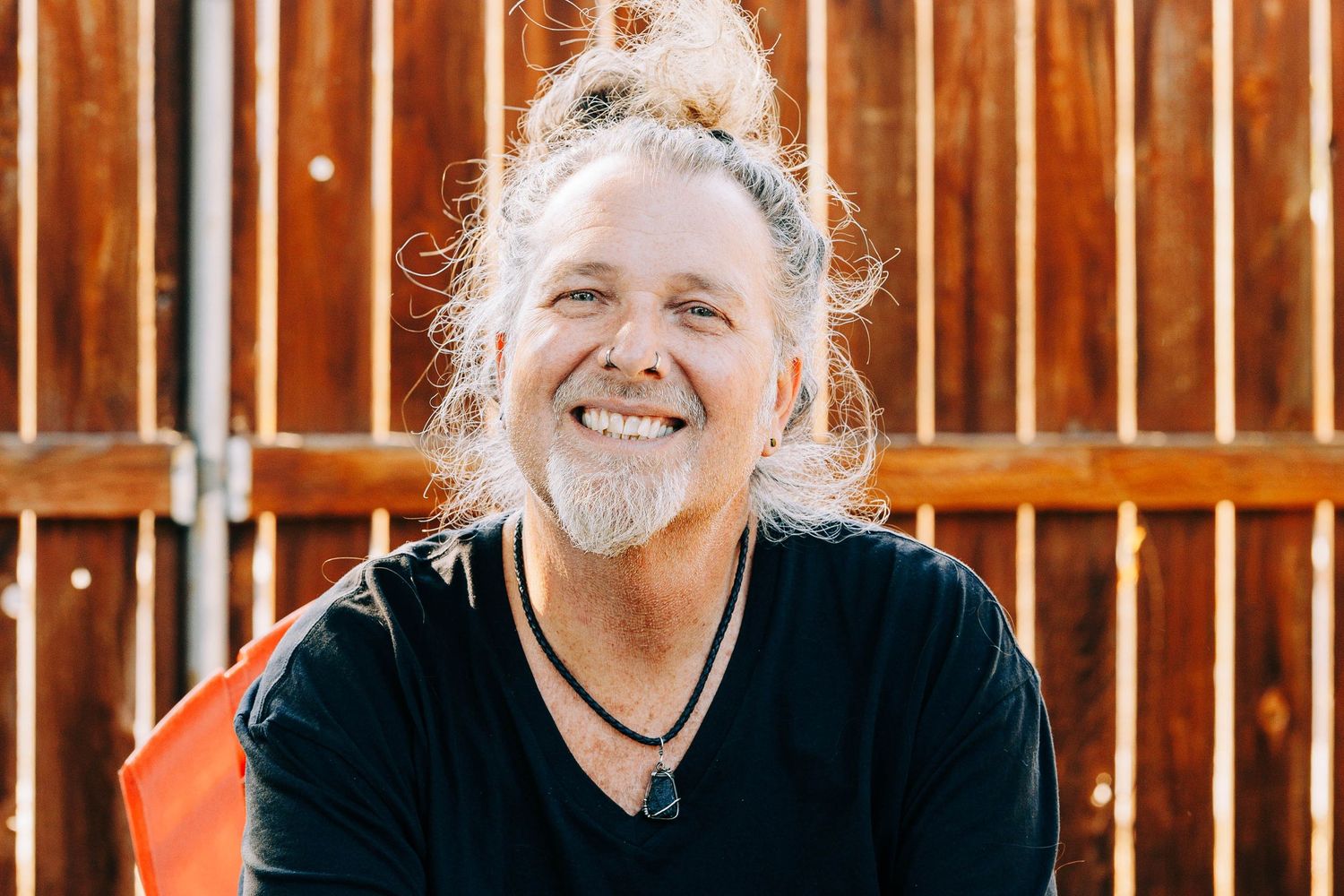 Smiling middle-aged man with long gray hair in a bun sitting against a rustic wooden fence outdoors.