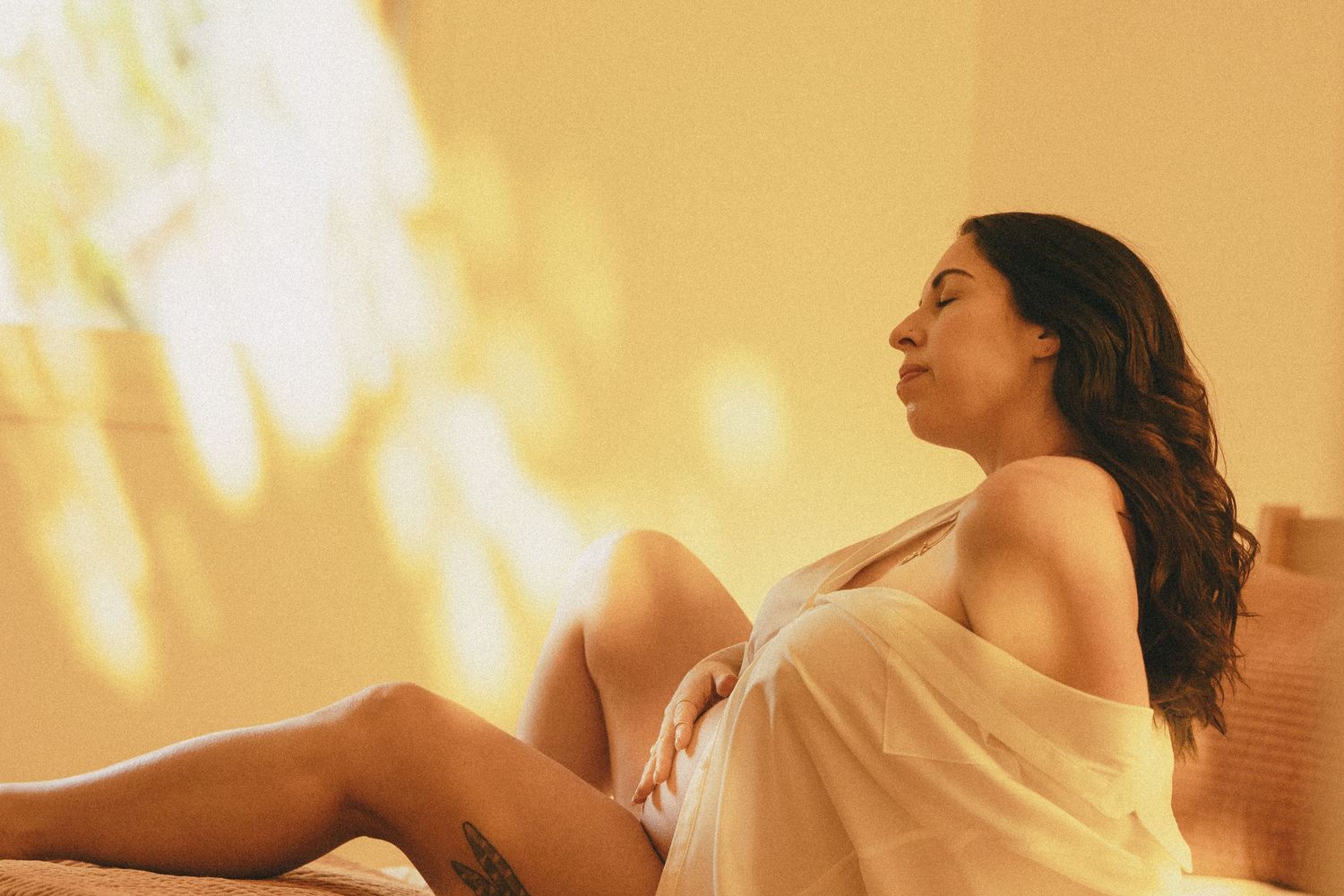 Pregnant woman draped in sheer fabric sitting on bed with eyes closed in warm amber glowing light.