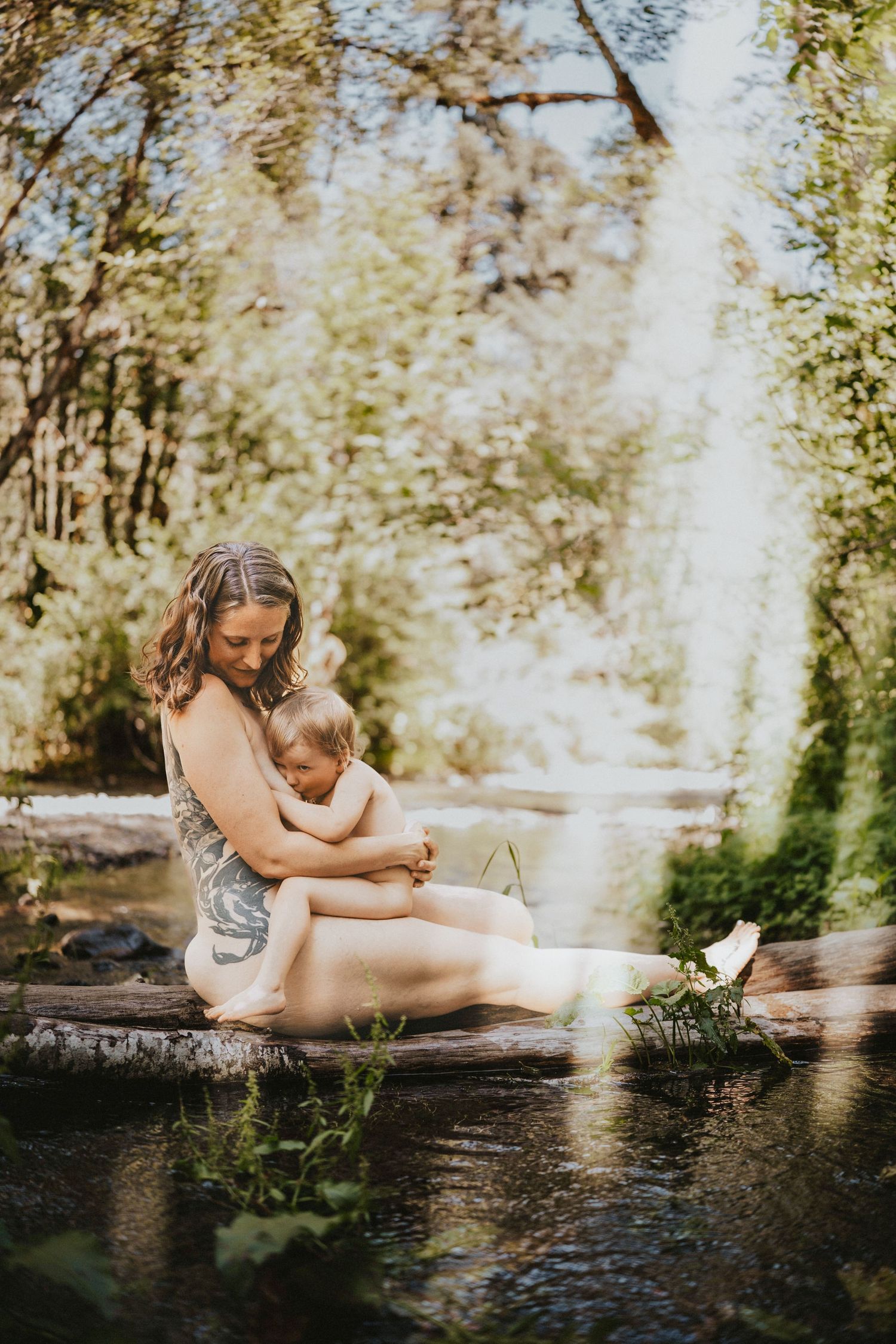A mother sits on a log by a peaceful creek, tenderly holding her baby in a lush green outdoor setting.