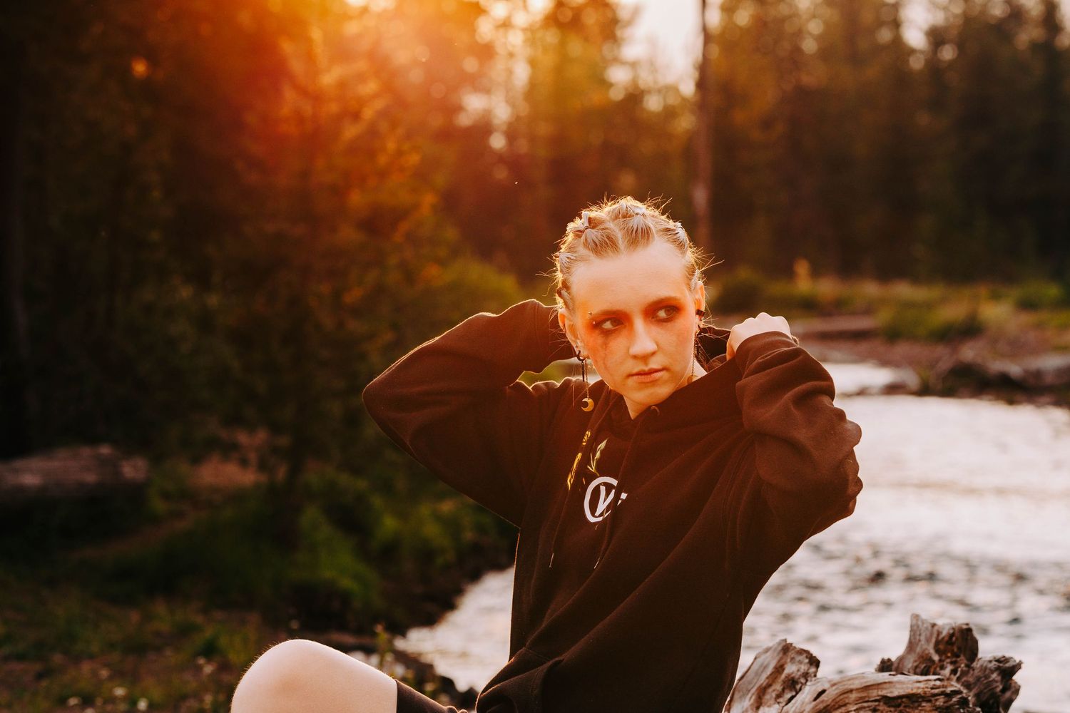 Young woman in black hoodie poses on log with golden sunset light in forest setting.