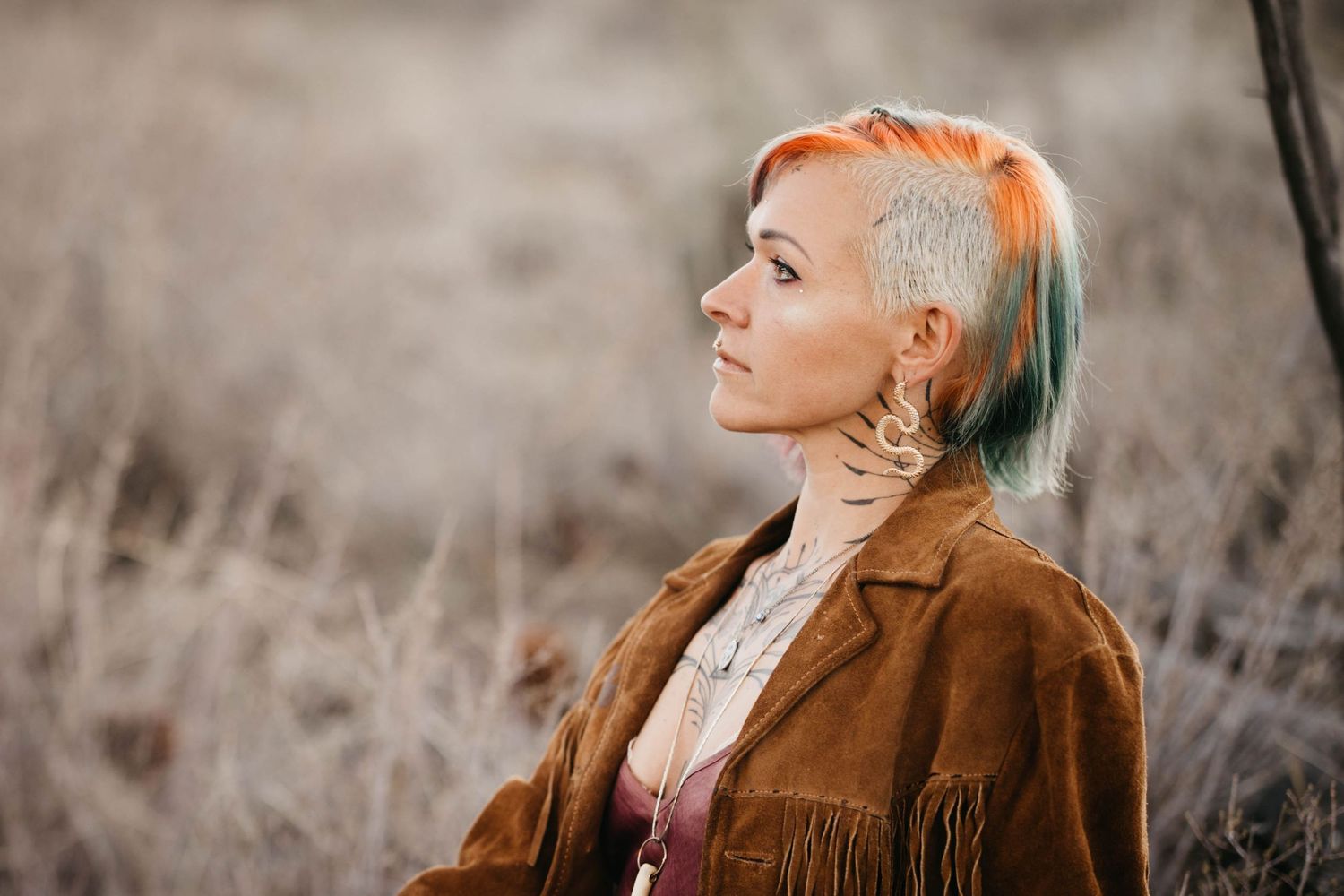 Woman with short multicolored hair wearing a fringed suede jacket standing in a dry field.