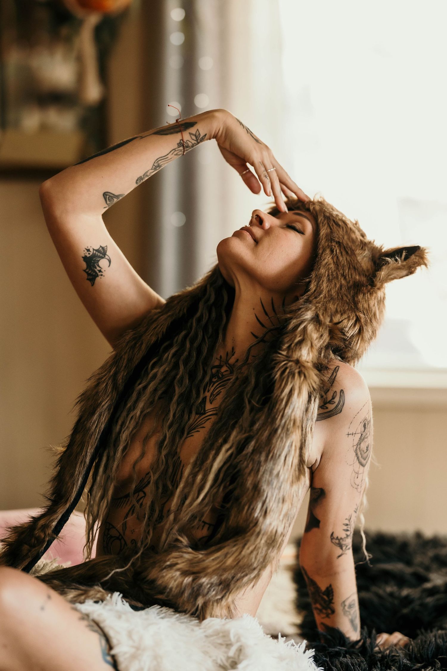 Tattooed woman wearing a fur wolf hood poses dramatically with eyes closed and arm raised overhead.