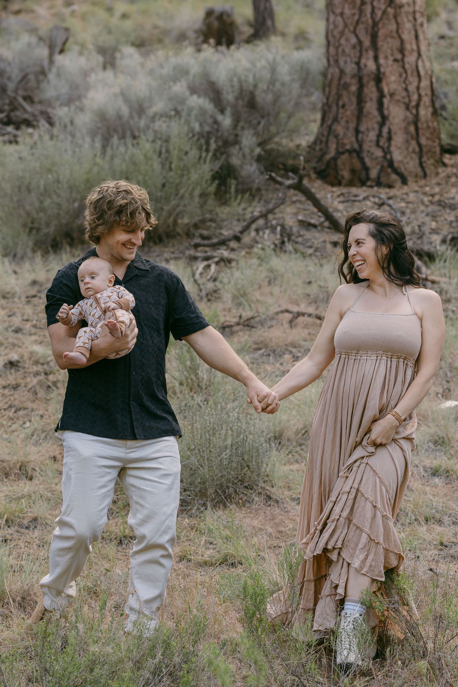 Young couple holding hands and carrying newborn baby while walking through natural desert brush landscape.