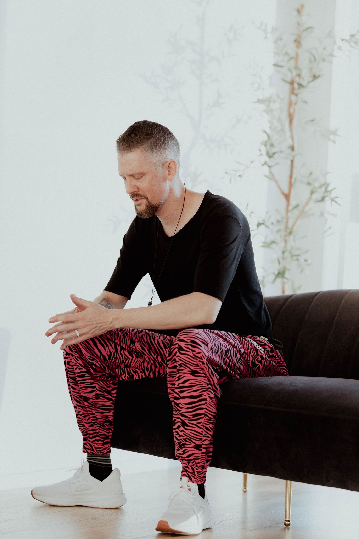 Man in black t-shirt and pink zebra print pants sitting on dark sofa in bright white room.