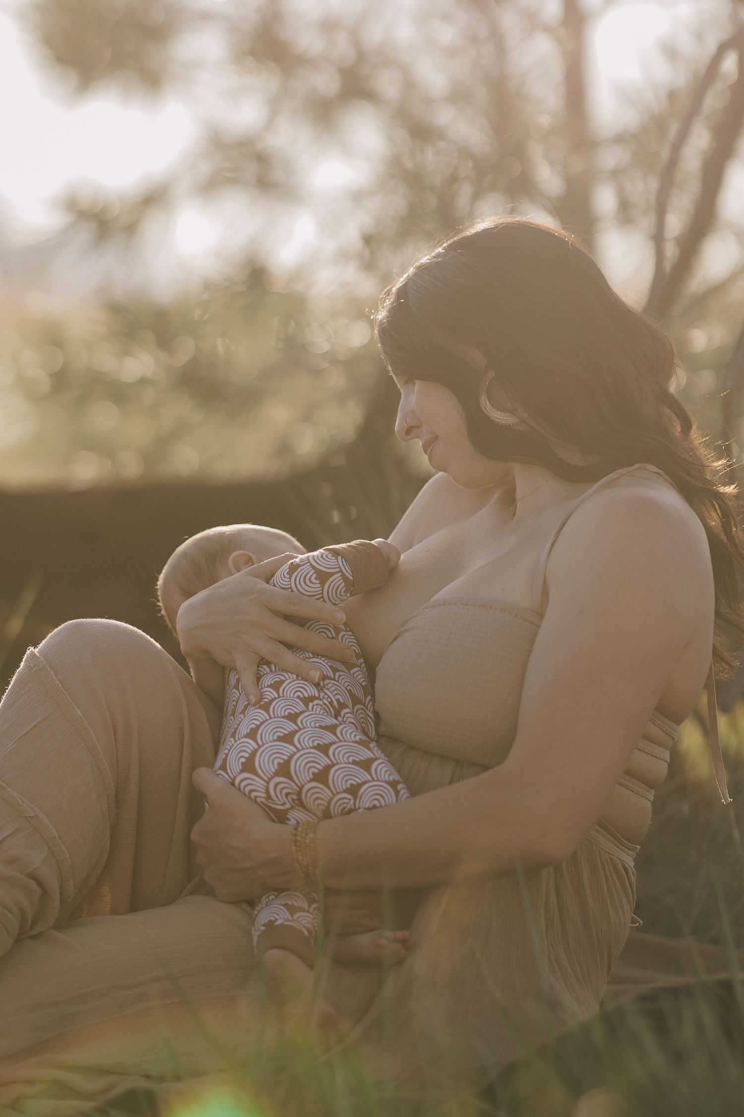 Mother breastfeeding newborn baby outdoors in warm golden sunlight during lifestyle photography session.