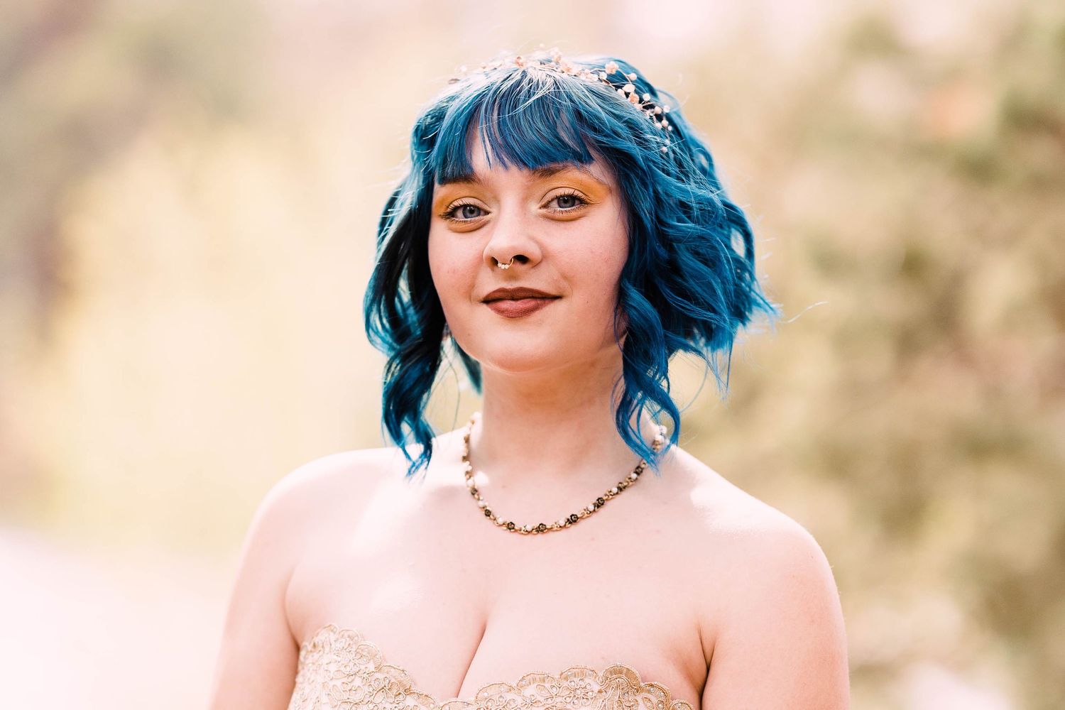 Young woman with blue wavy hair and floral crown wearing strapless lace gown and beaded necklace outdoors.