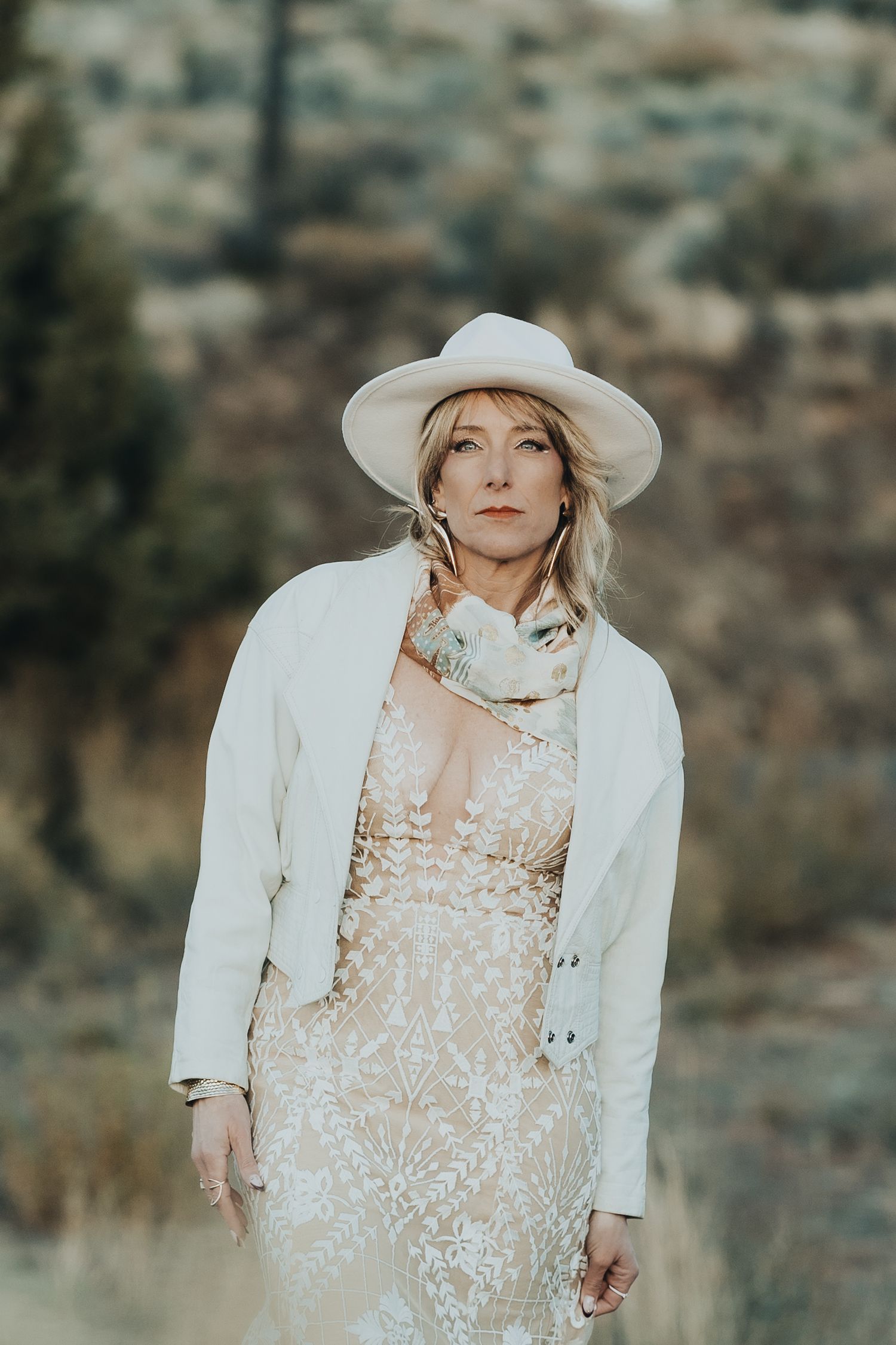 Blonde woman in white hat, lace dress, and white jacket posing in rustic outdoor landscape.