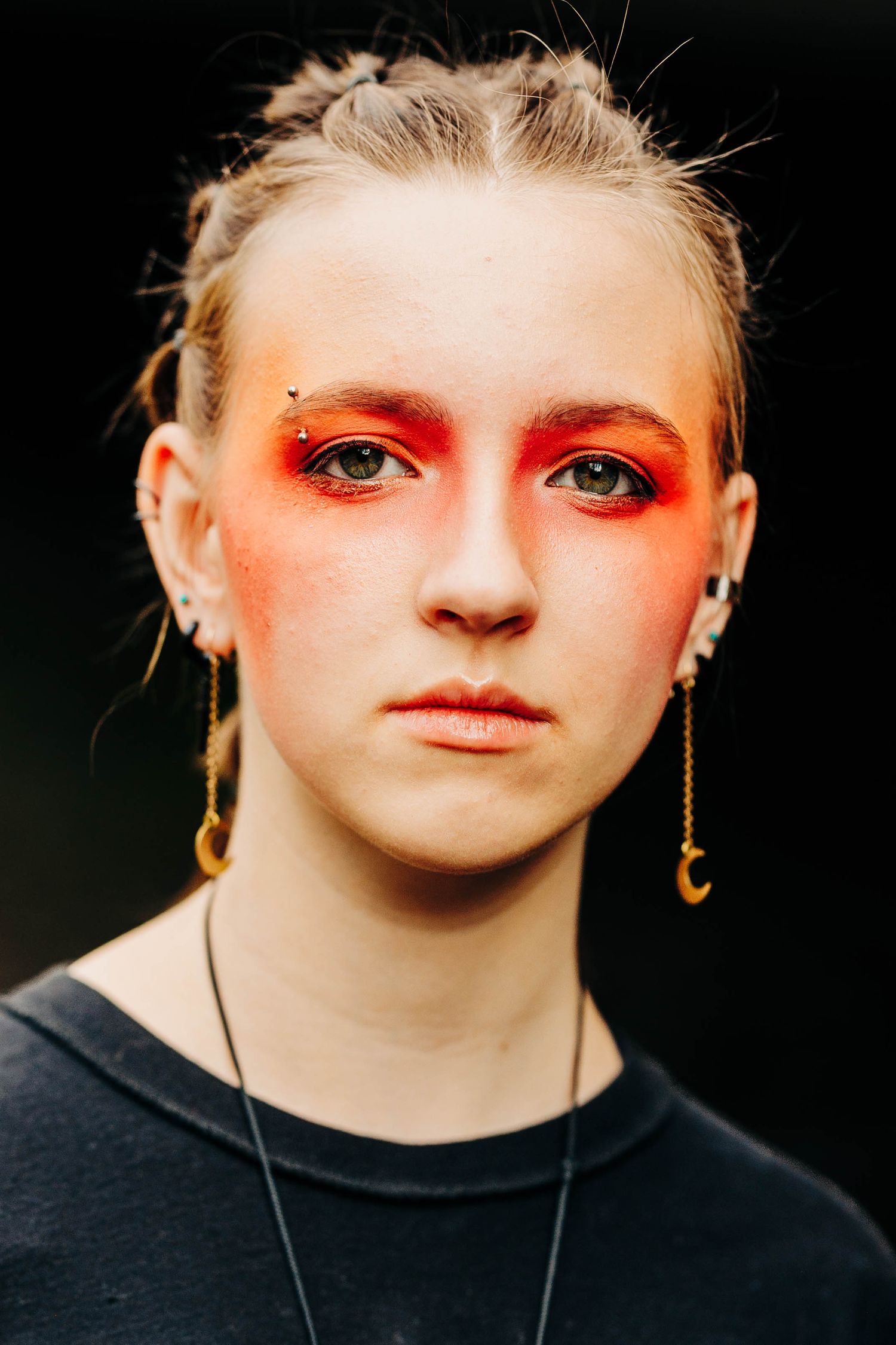 Close-up portrait of a young woman with dramatic orange-red eye makeup, moon earrings, and braided hair against dark background.