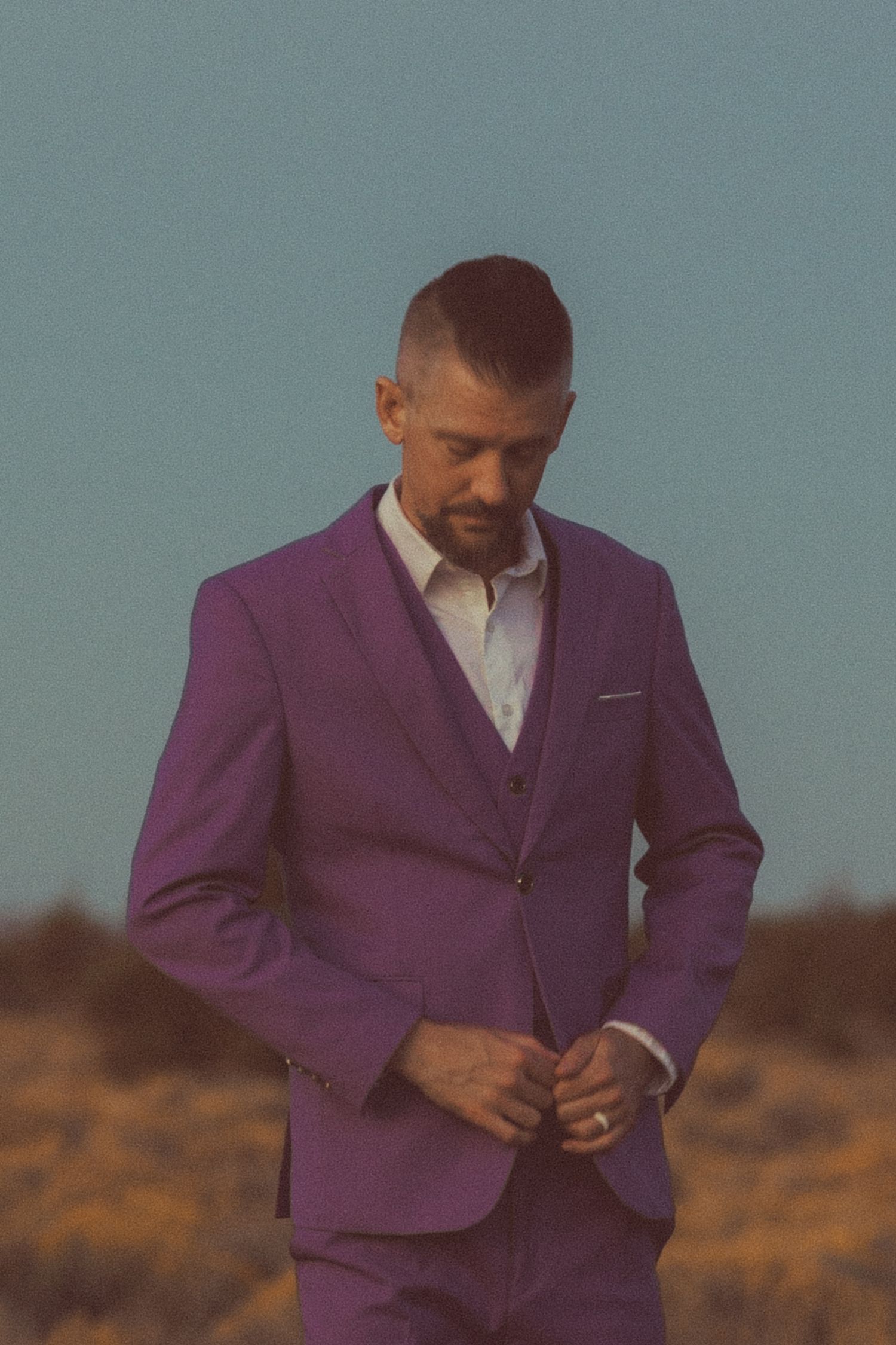 Man in purple three-piece suit looking down while buttoning jacket in desert setting.