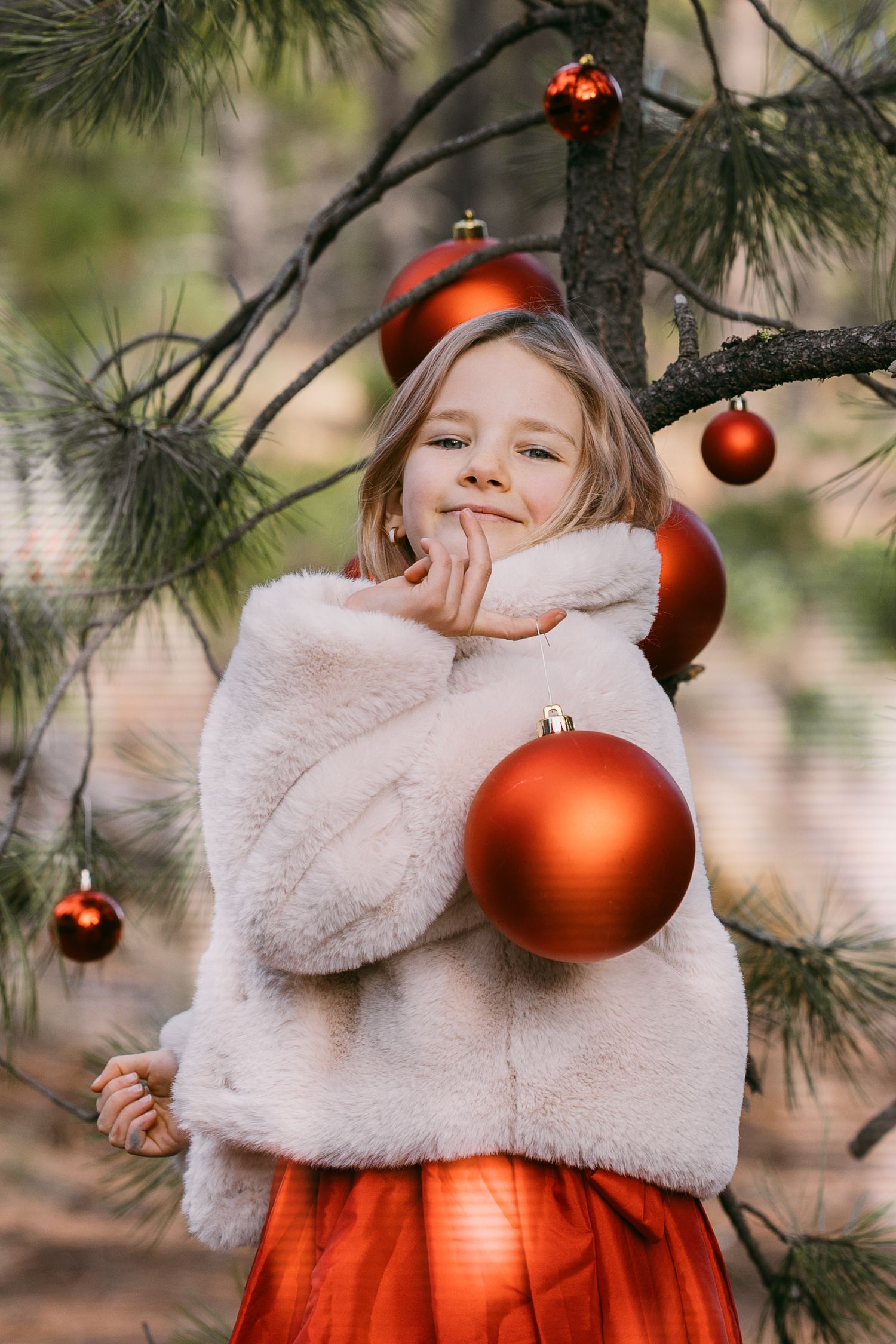 Cute blonde girl in white fur coat and red skirt hugging oversized red Christmas ball ornament outdoors.
