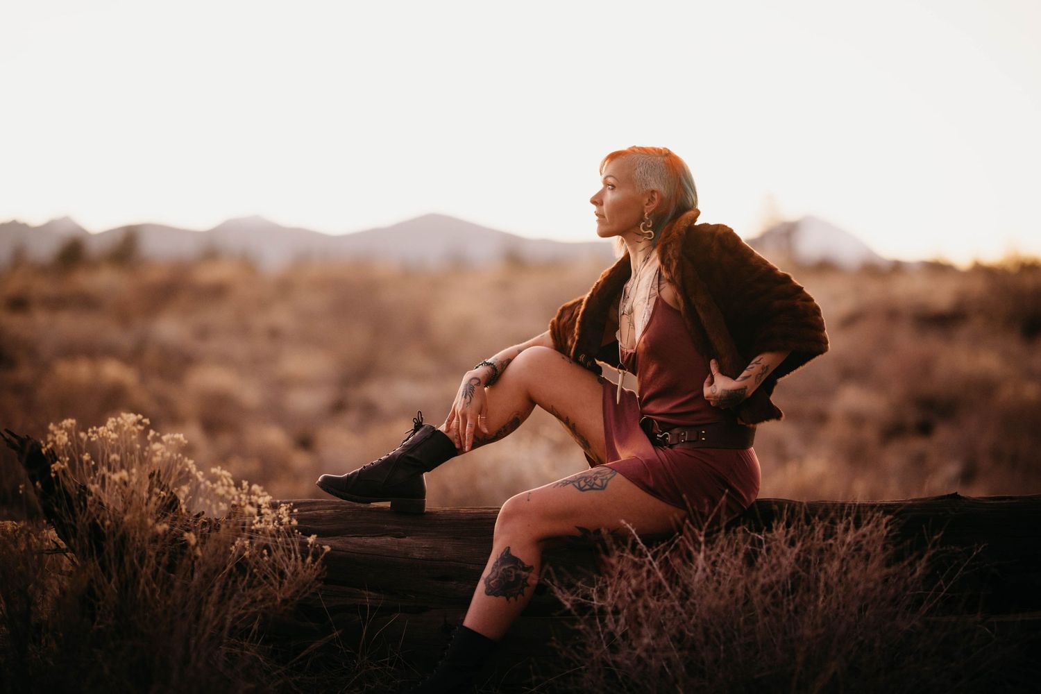 Woman in burgundy dress and black boots sits on log in golden desert landscape.