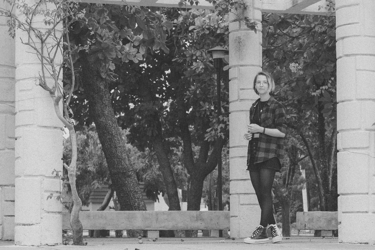Black and white photo of a woman standing between stone columns in a tree-lined park.