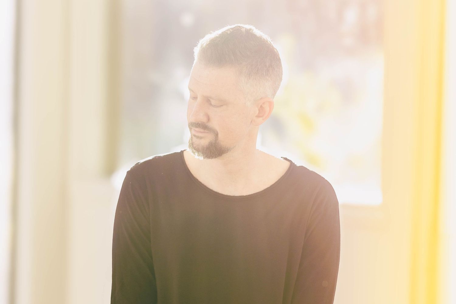Bearded man in black long-sleeve shirt with eyes closed in soft bright natural light.