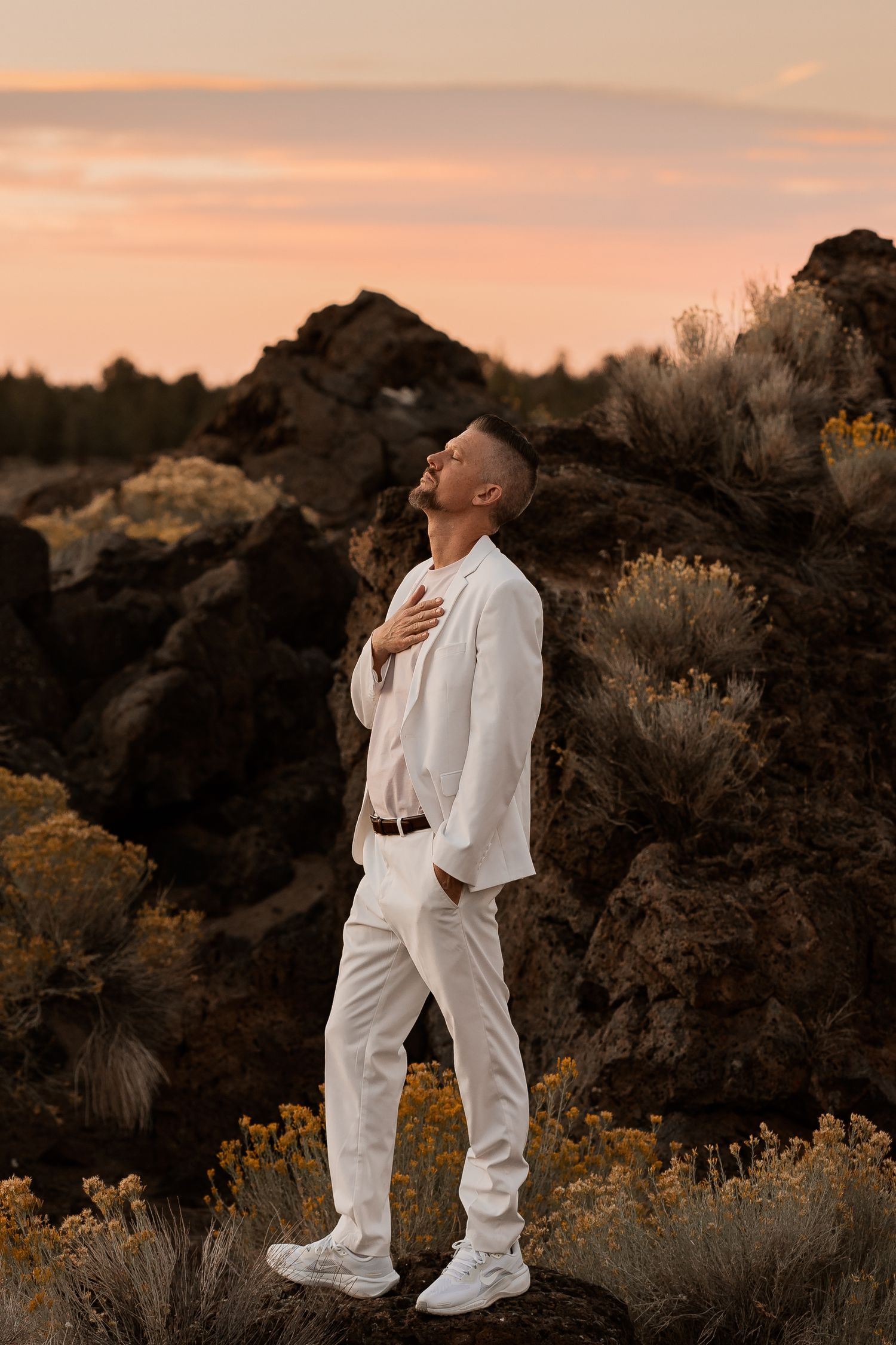 Man in white suit poses on volcanic rocks at sunset with sagebrush and wildflowers in background.