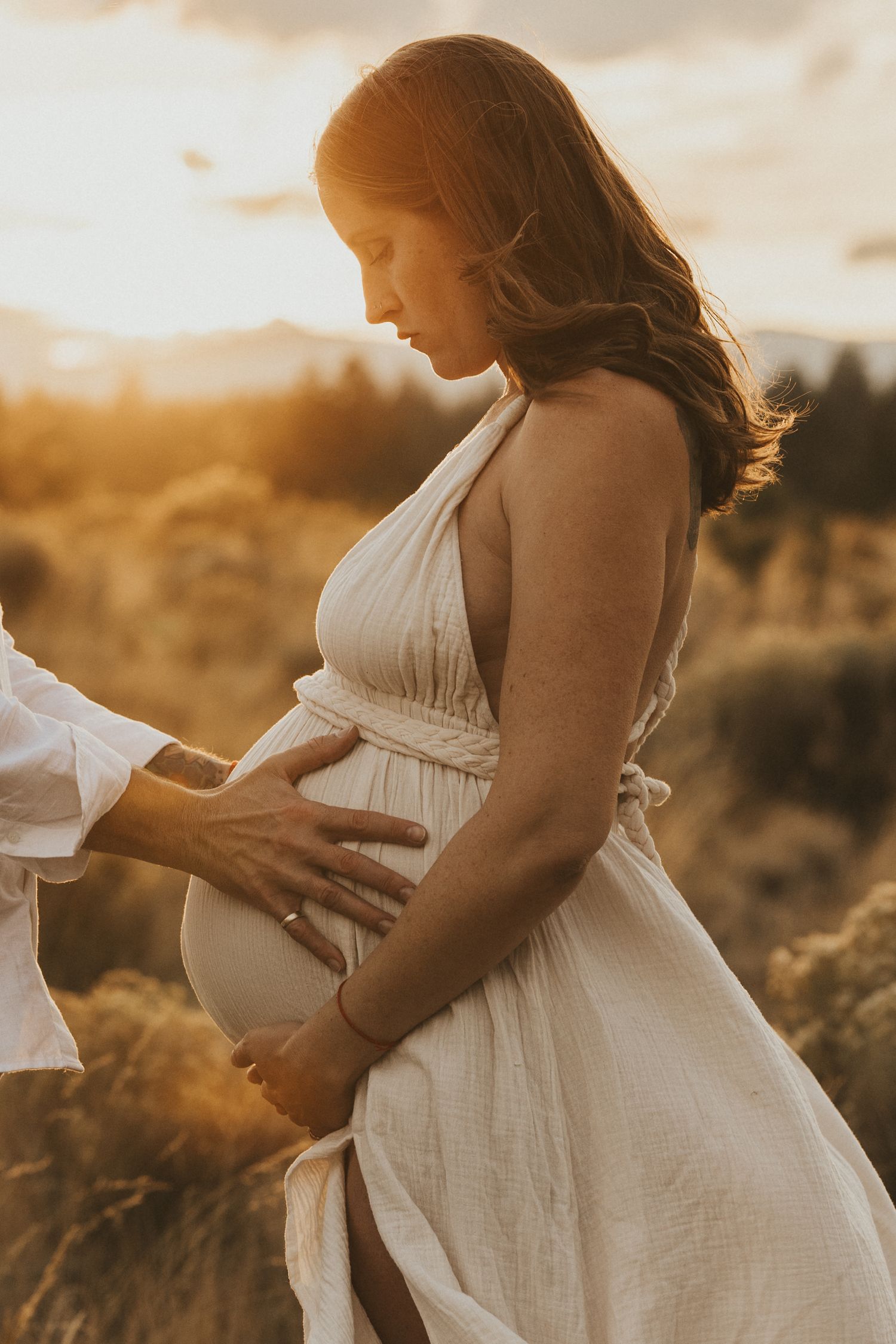 Golden sunset maternity photo of pregnant woman in white dress with partner's hands on her belly.