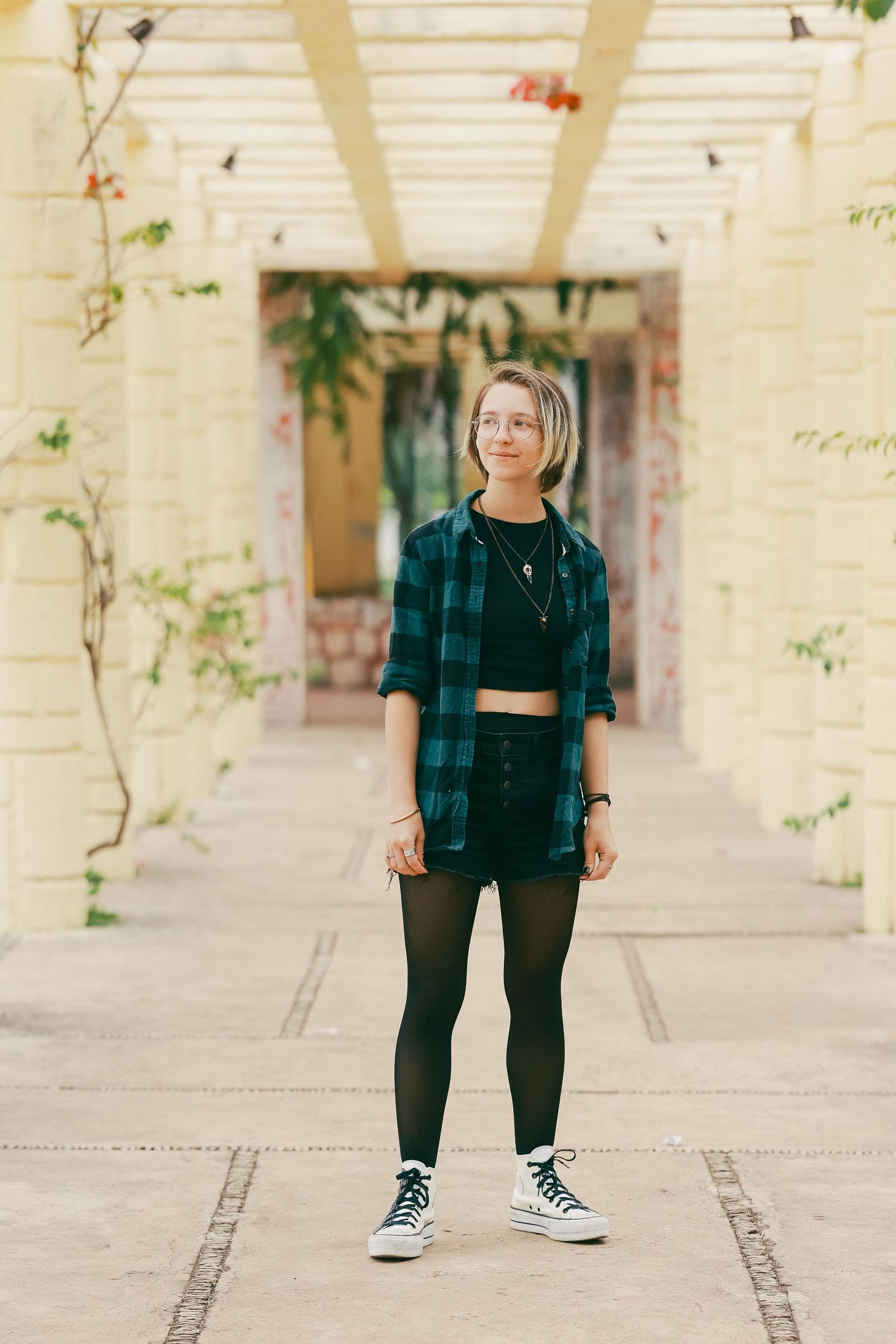 A young woman in plaid shirt, black skirt, and tights stands in a yellow pergola walkway.