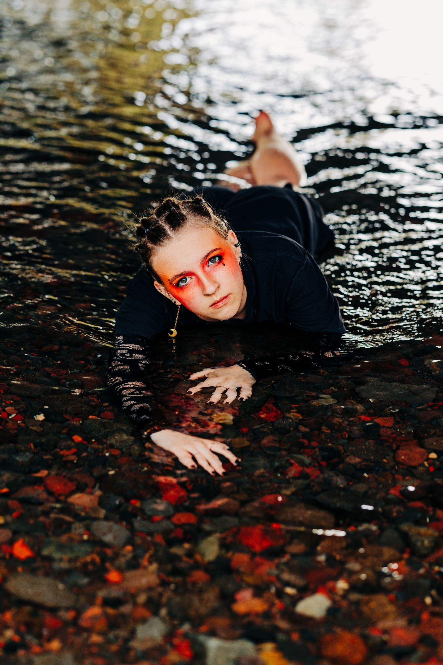Young woman with bold red eye makeup lies in shallow rocky water wearing black outfit.