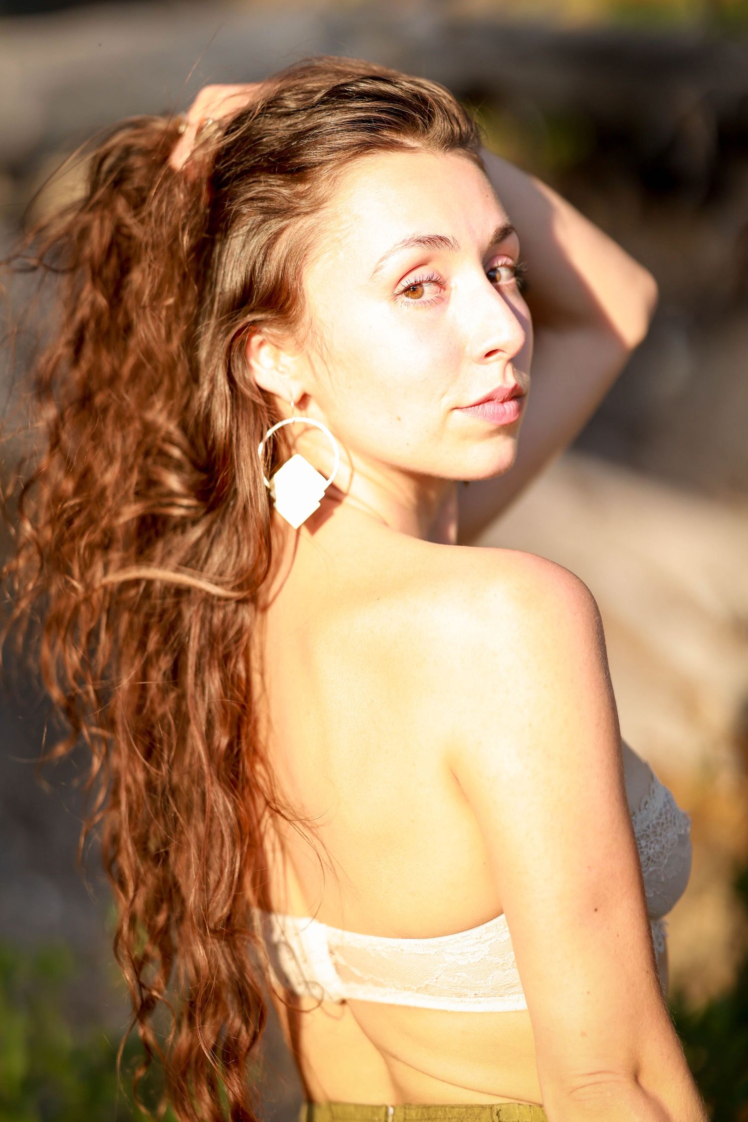 Young woman with long curly auburn hair poses outdoors in warm sunlight wearing large geometric earrings.