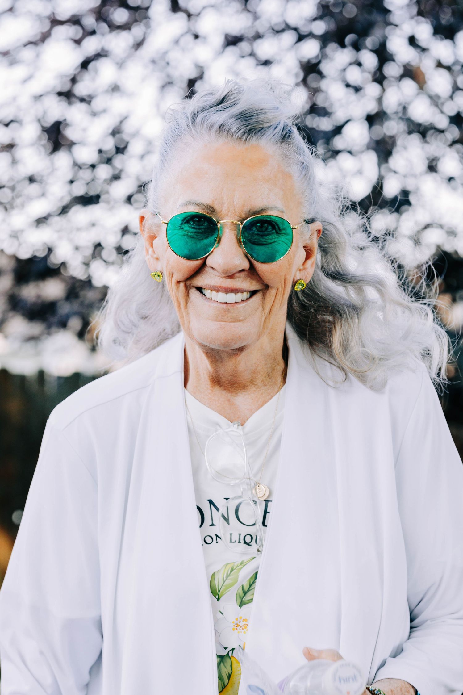 Elderly woman with silver hair smiling in white outfit and teal reflective sunglasses.
