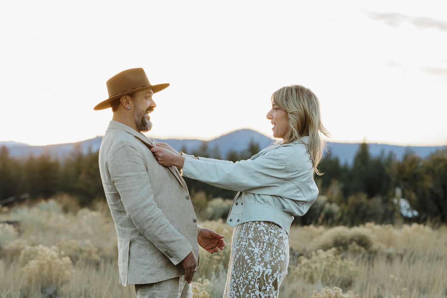 Boho couple sharing a playful moment in an open field with mountain views at sunset.