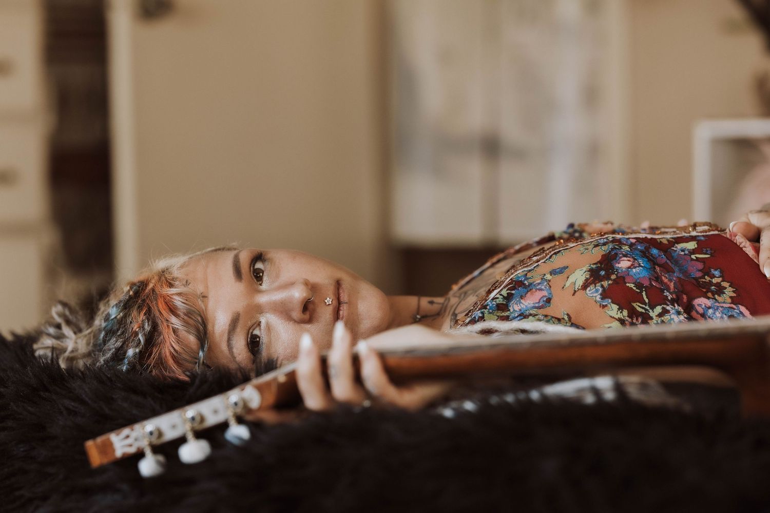 Tattooed woman with short hair lying down holding a guitar neck in a cozy indoor setting.