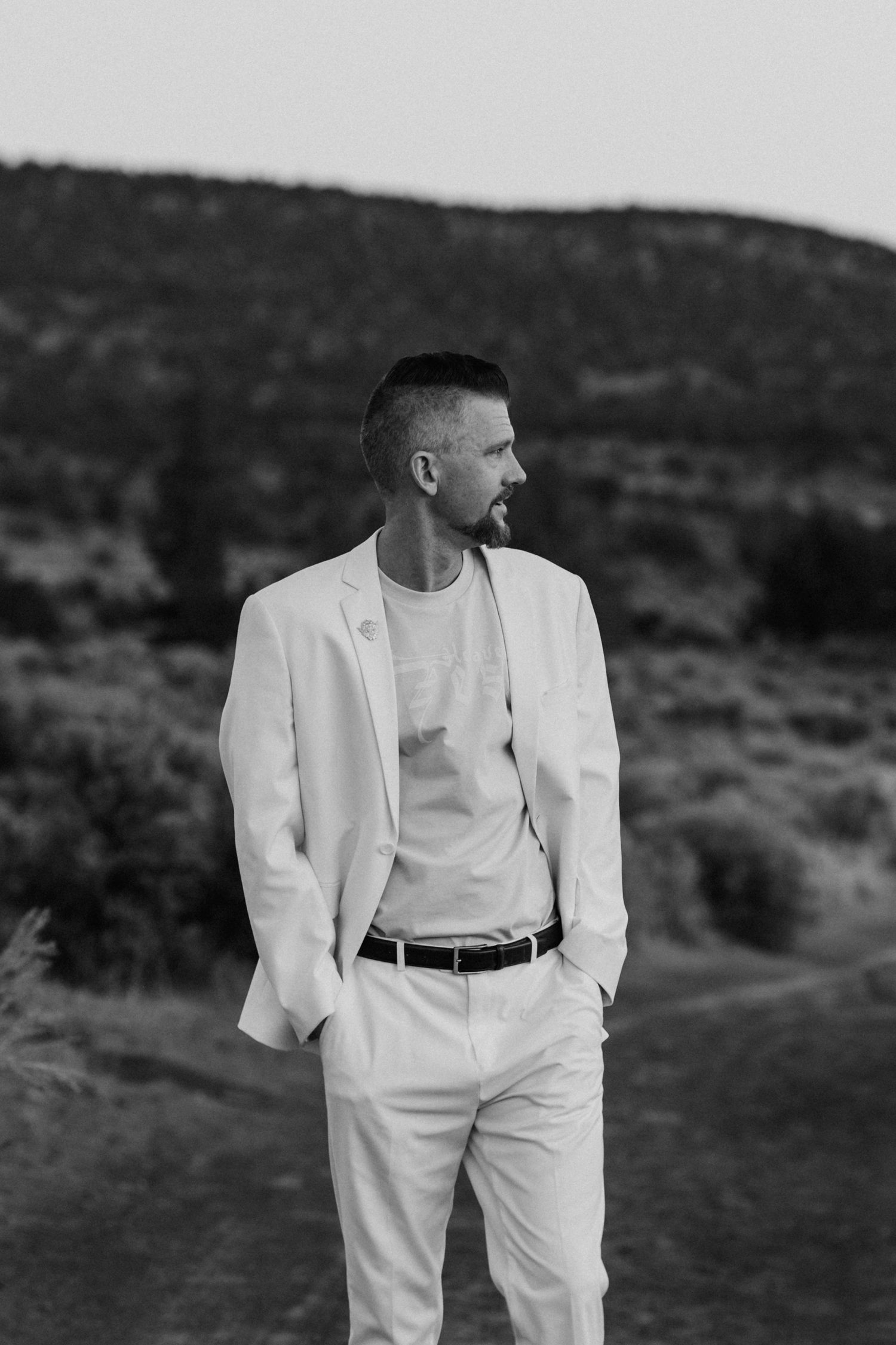 Man in white blazer and pants standing outdoors in black and white photo with hills behind.