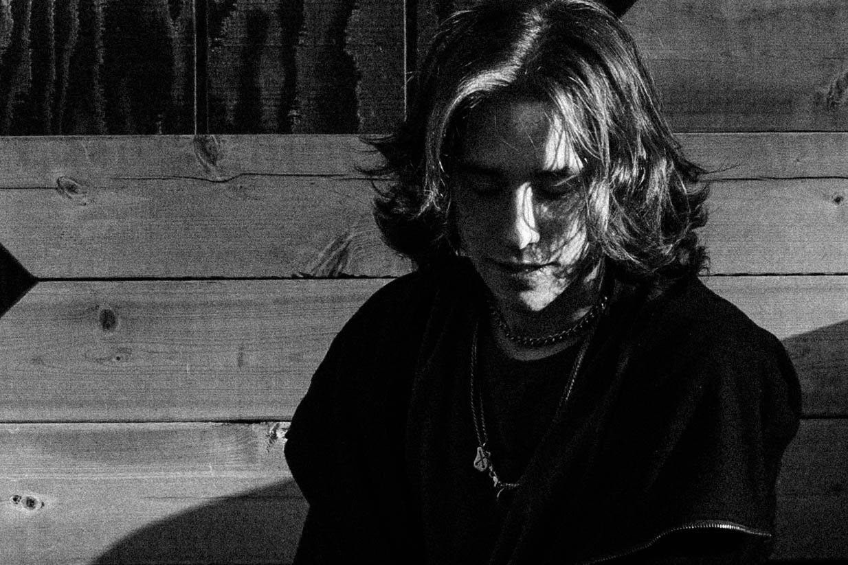 Black and white portrait of a young person with long hair looking down near a wooden wall.