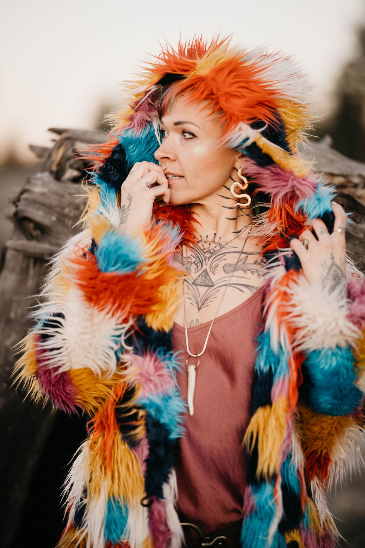 Woman in colorful multicolored faux fur hooded coat with chest tattoos poses outdoors.