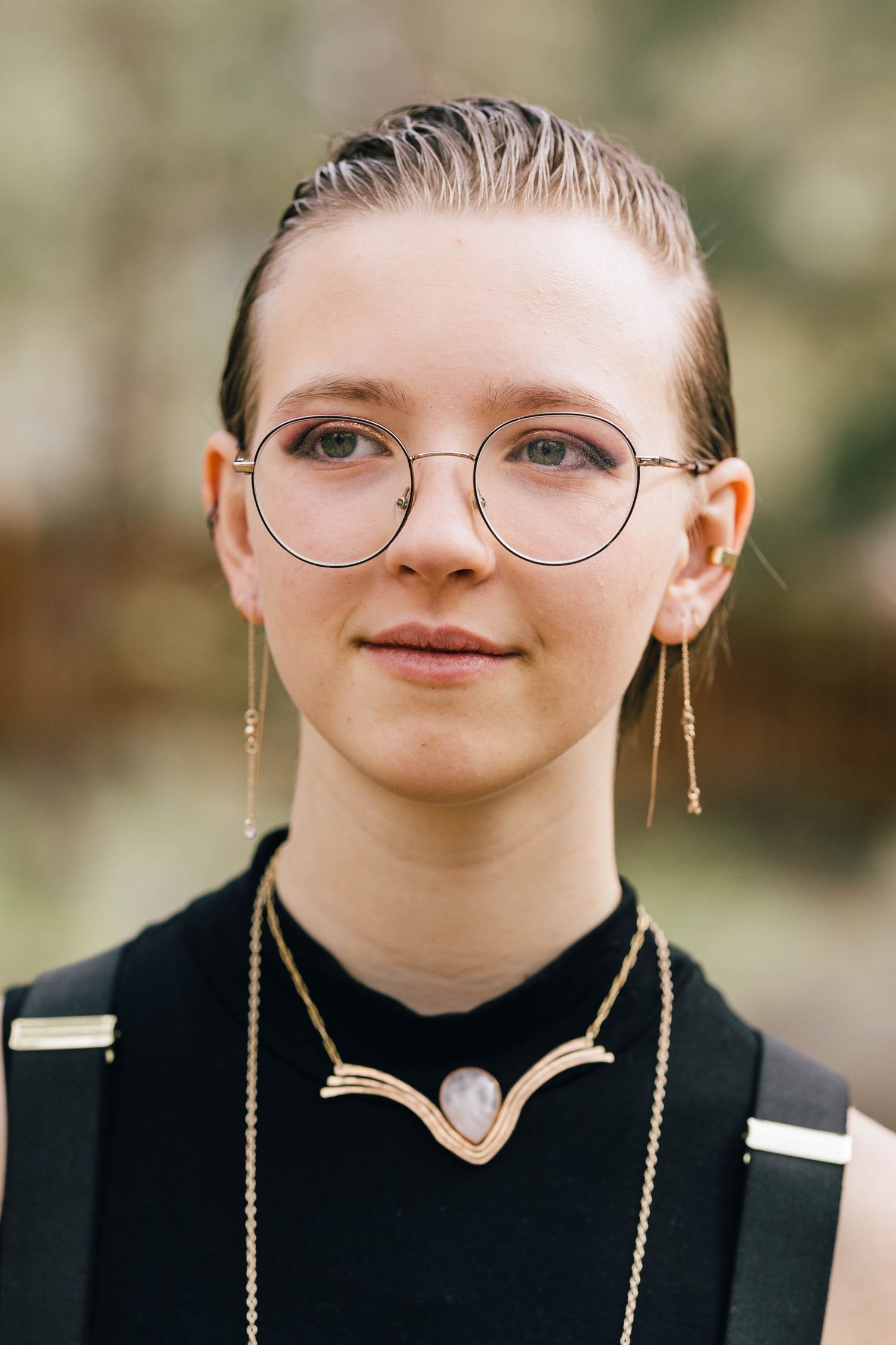 Close-up portrait of young person with round glasses, dangling gold earrings, and crescent moon necklace outdoors.