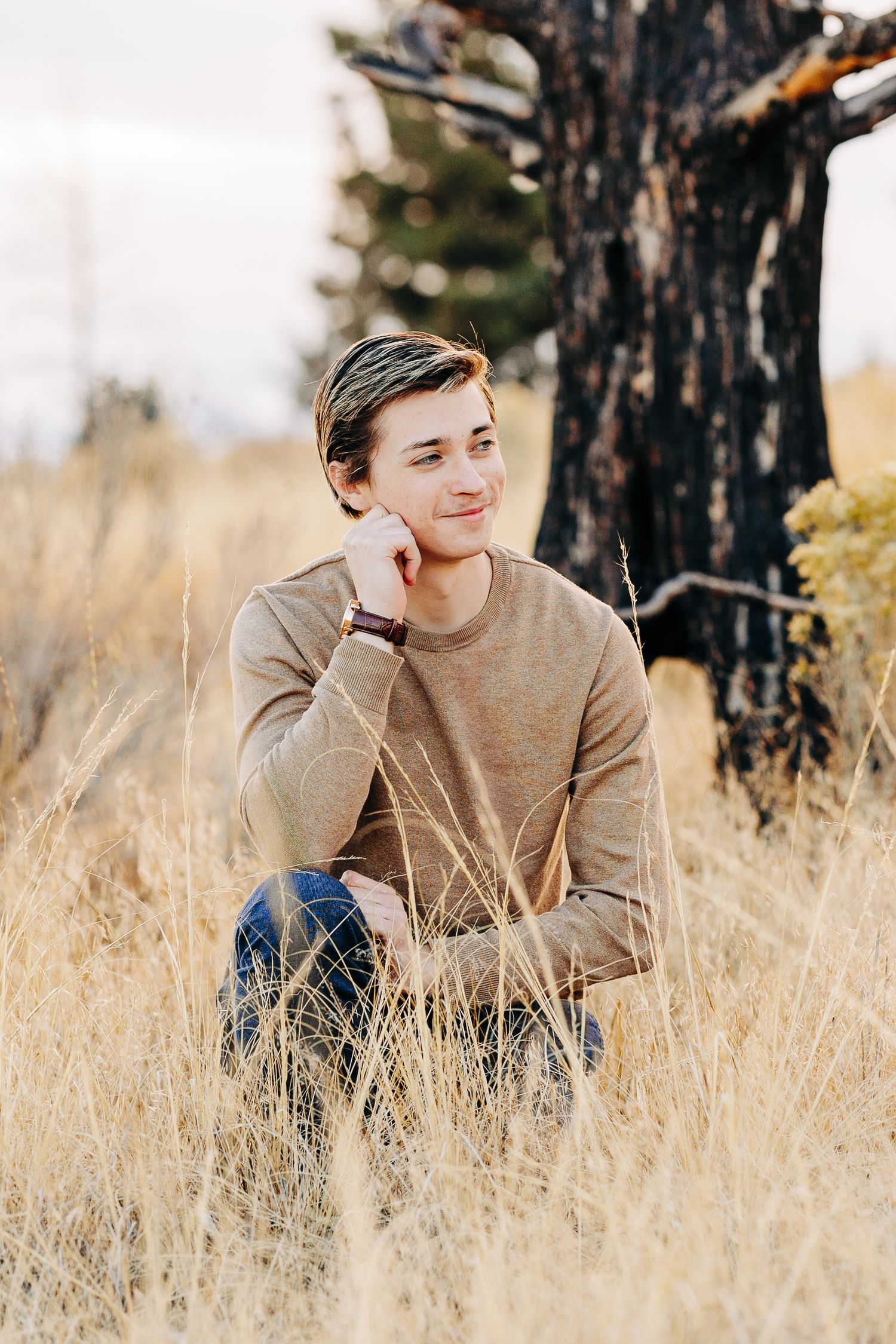 Young man in beige sweater sitting in golden dry grass field leaning against a tree outdoors.