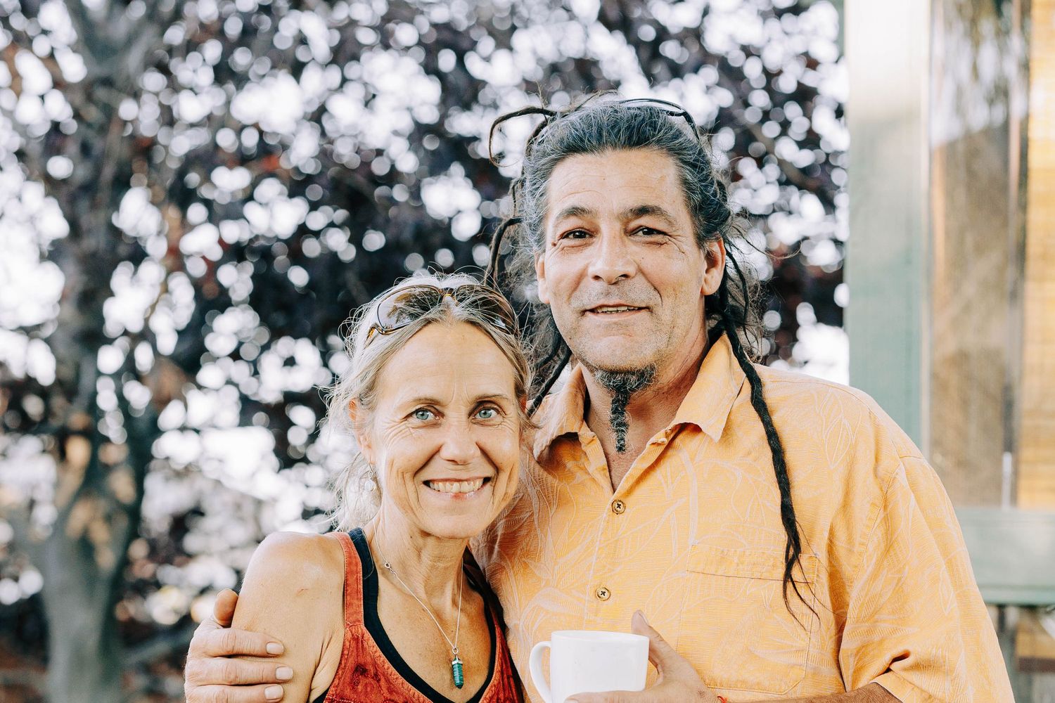 Smiling couple embracing outdoors with man holding a cup near blooming trees in soft light.
