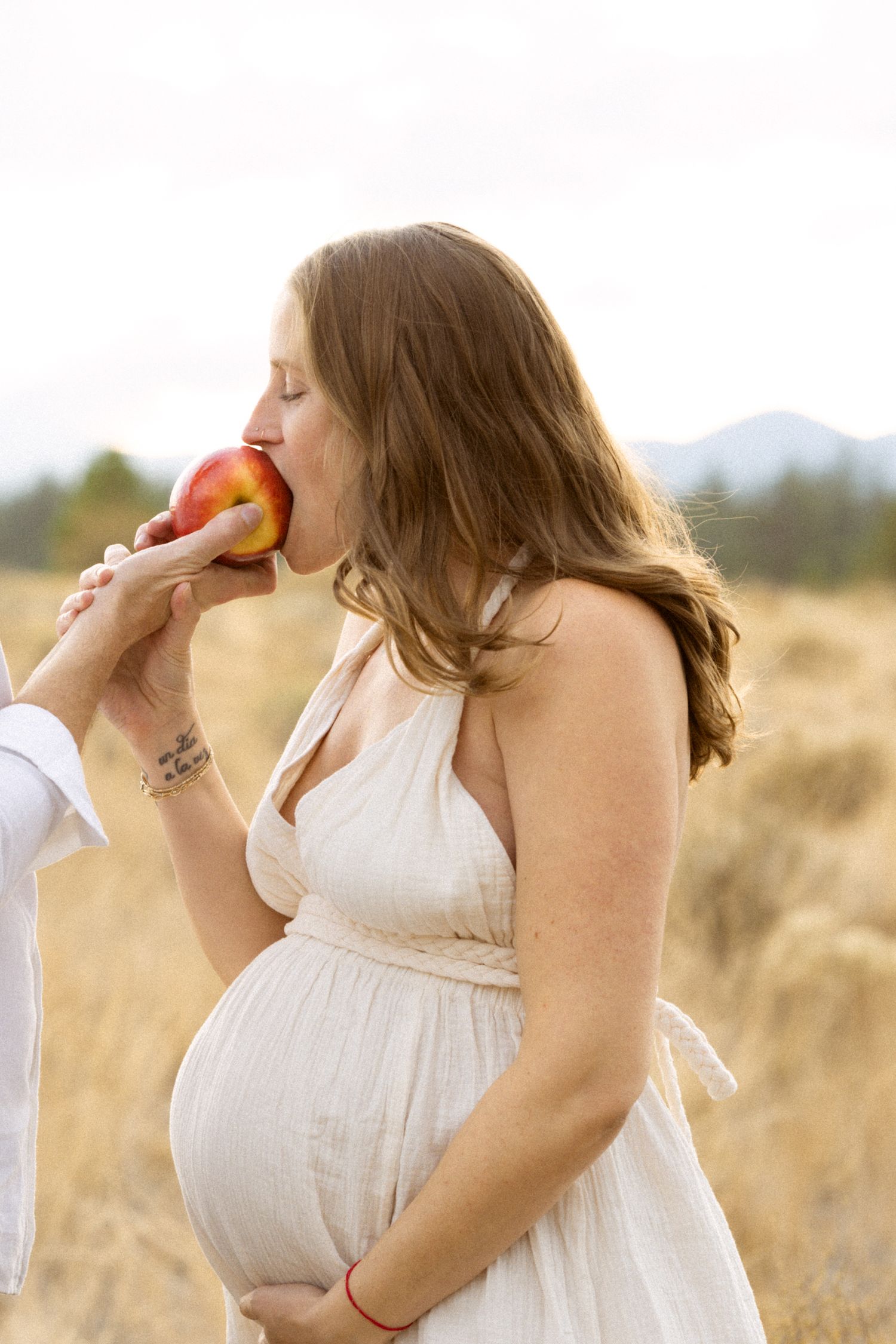 Pregnant woman in white dress biting a red apple in a golden field during maternity photo shoot.