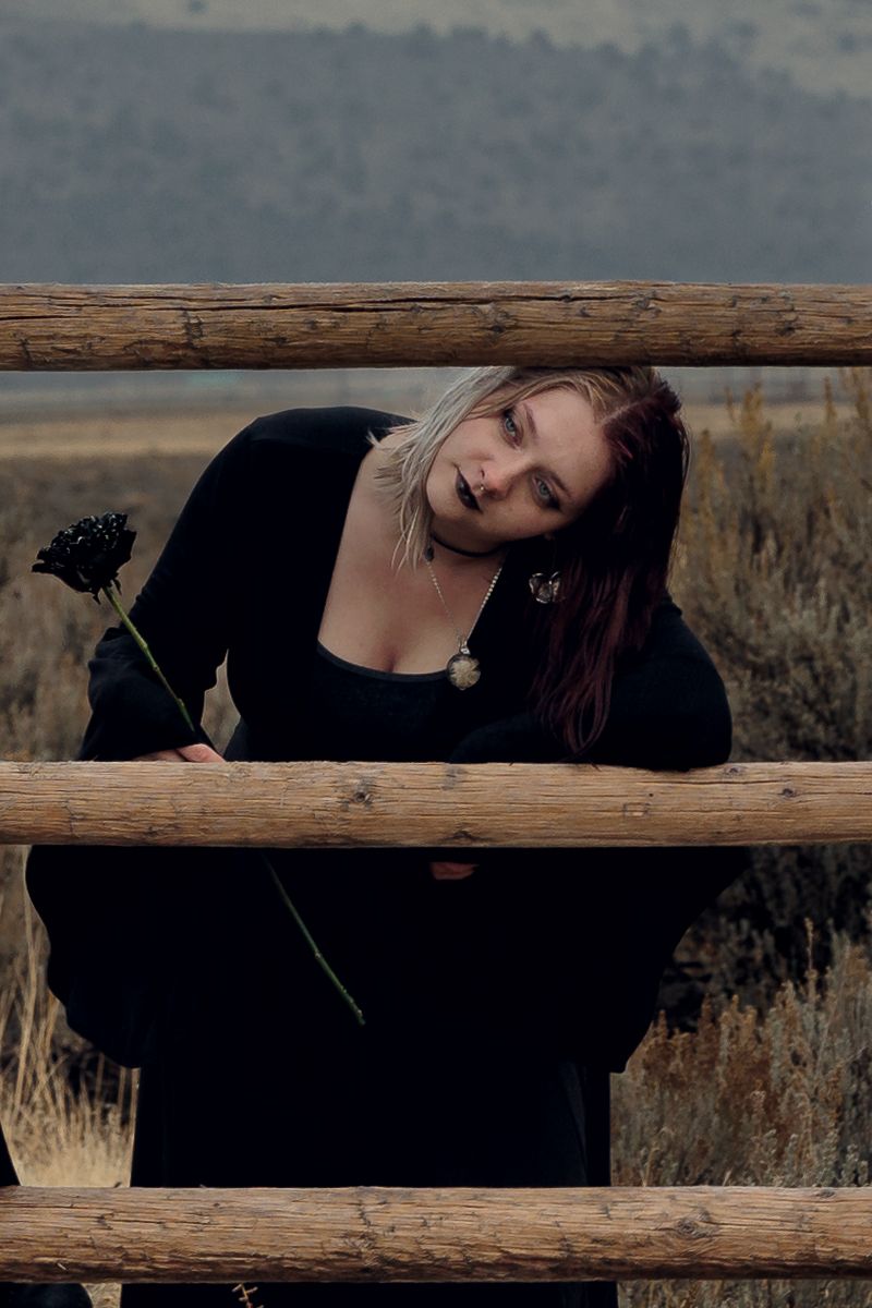 A blonde woman in a black dress leans through wooden fence rails holding a dark flower in a moody outdoor setting.