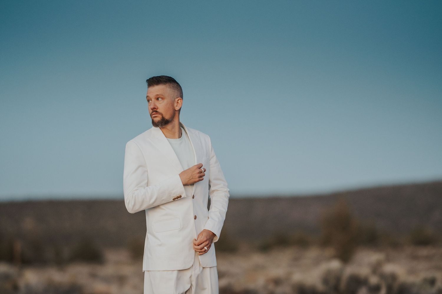 Man in white linen suit standing in desert landscape under clear blue sky.