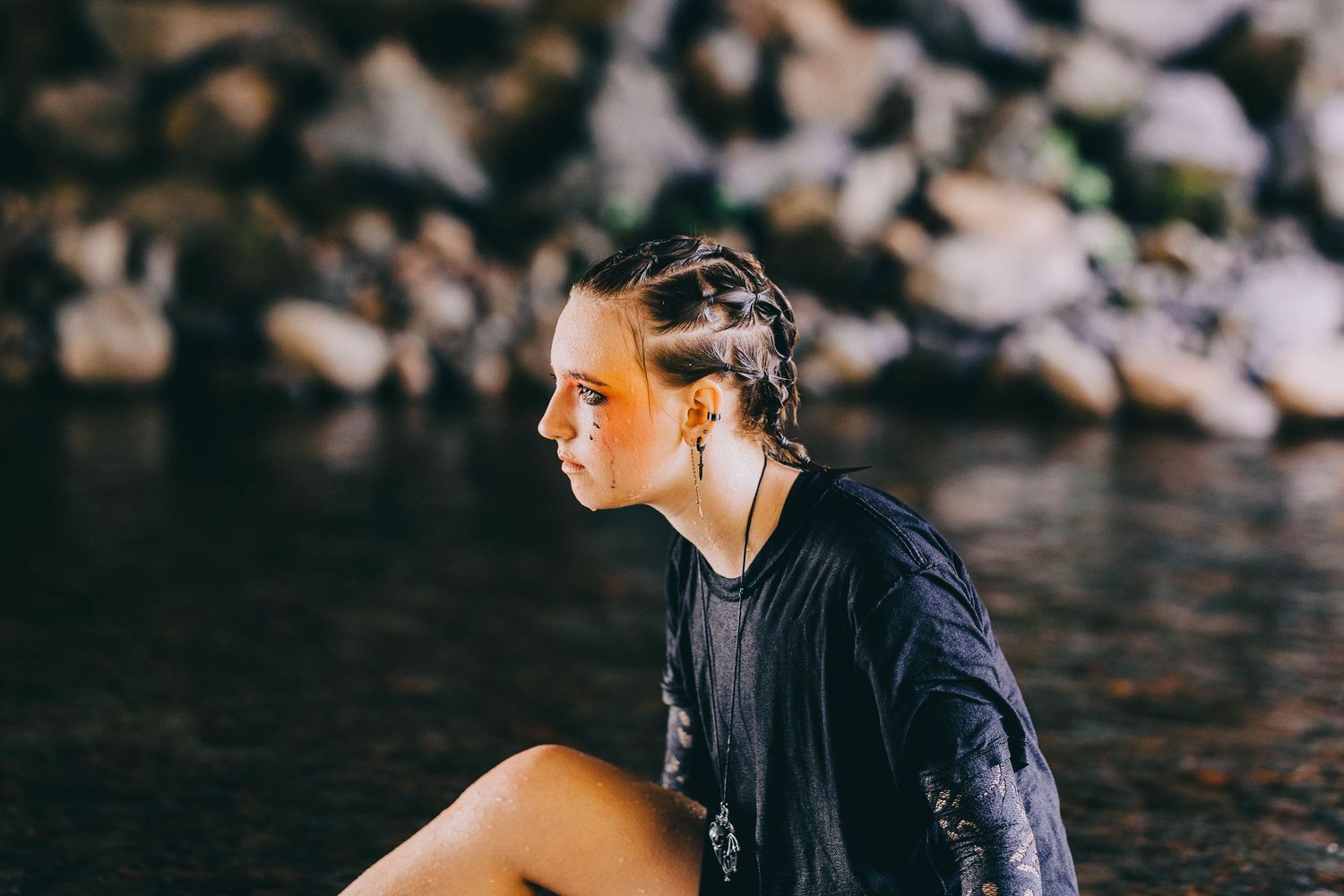 Young woman with braided hair and subtle makeup sits by rocky waterside in black top.