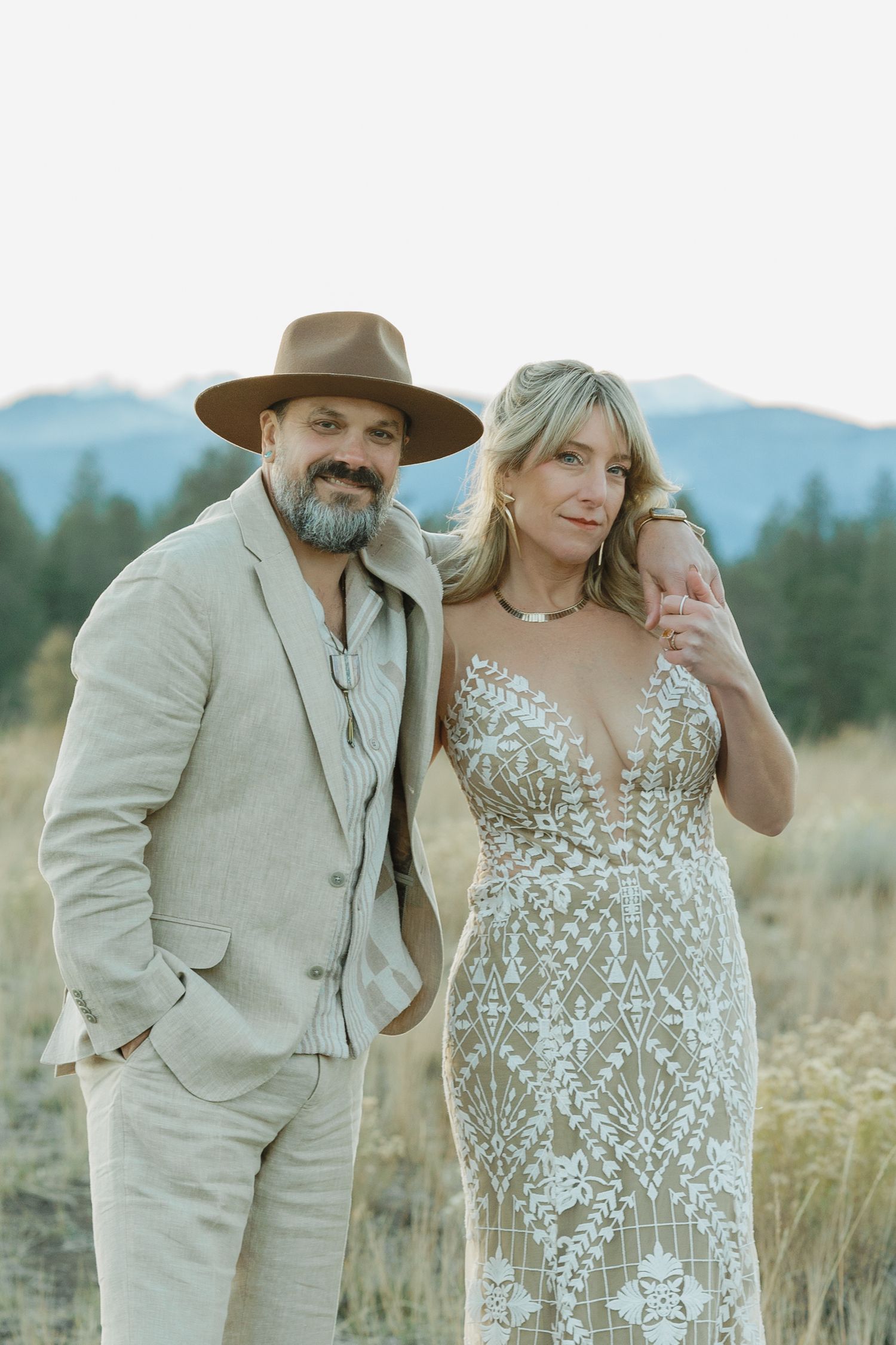 Boho wedding couple poses in field at dusk, groom in tan suit and wide-brim hat.