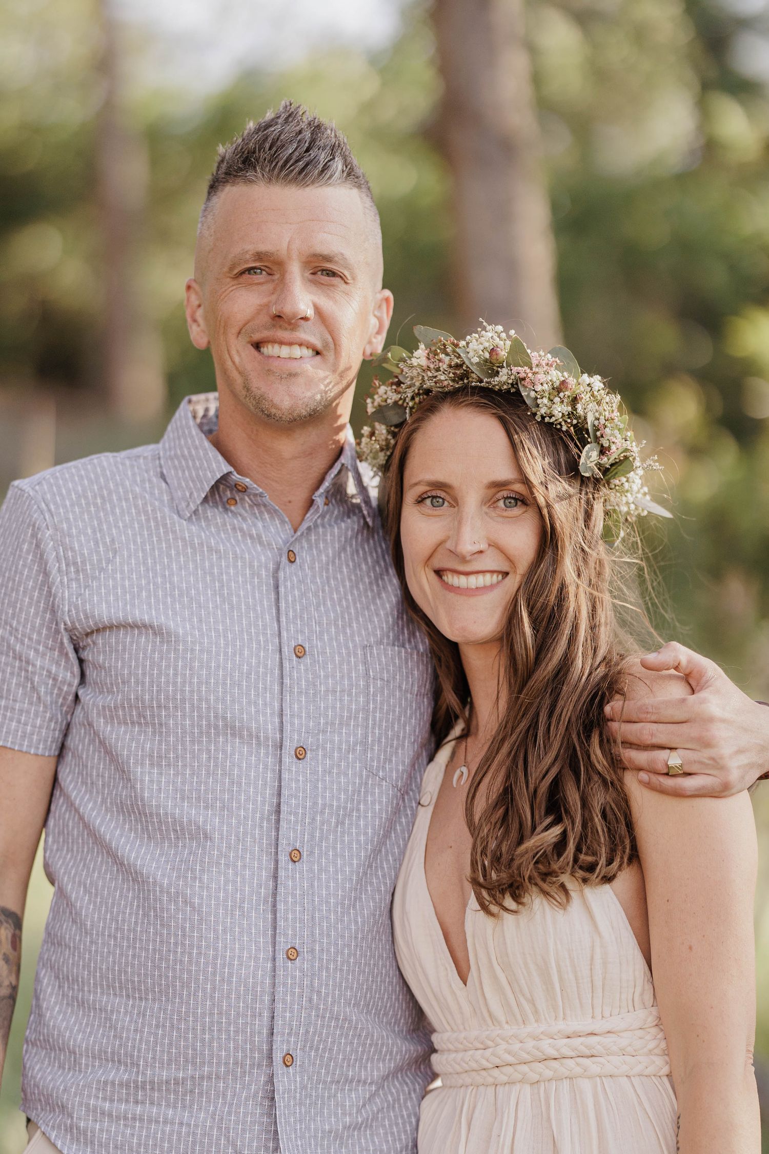 Smiling couple posing together outdoors, woman wearing floral crown and white dress.