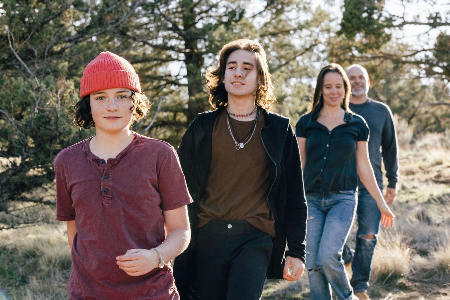 Four people walking outdoors through a natural landscape with trees in the background on a sunny day.