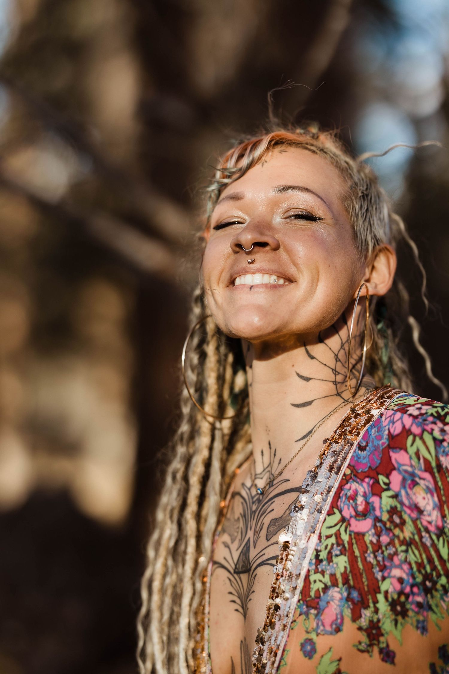 Smiling tattooed woman with dreadlocks and chest tattoos enjoying sunlight in a forest setting.