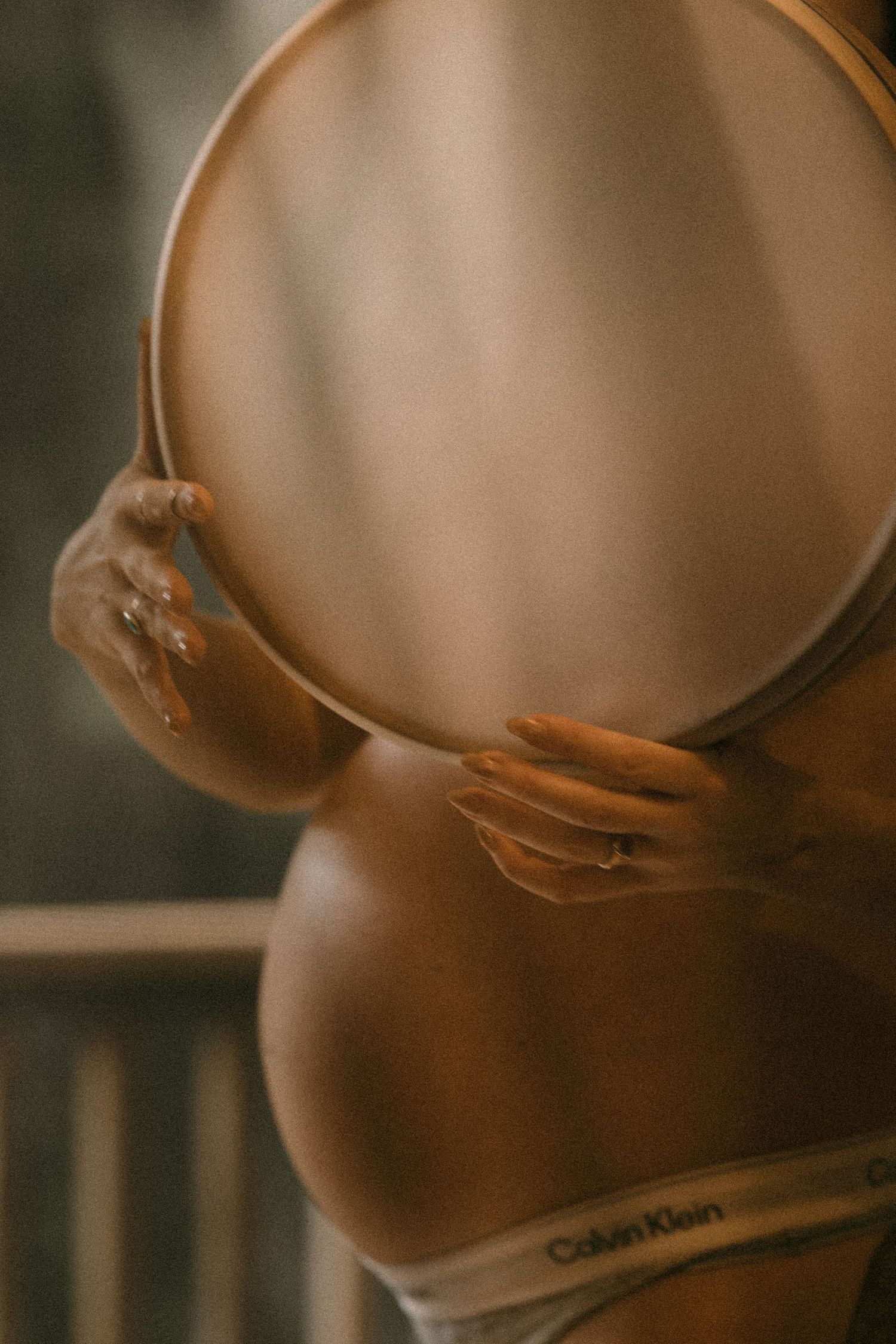 Side view of pregnant woman in Calvin Klein underwear holding a round mirror beside her glowing baby bump.