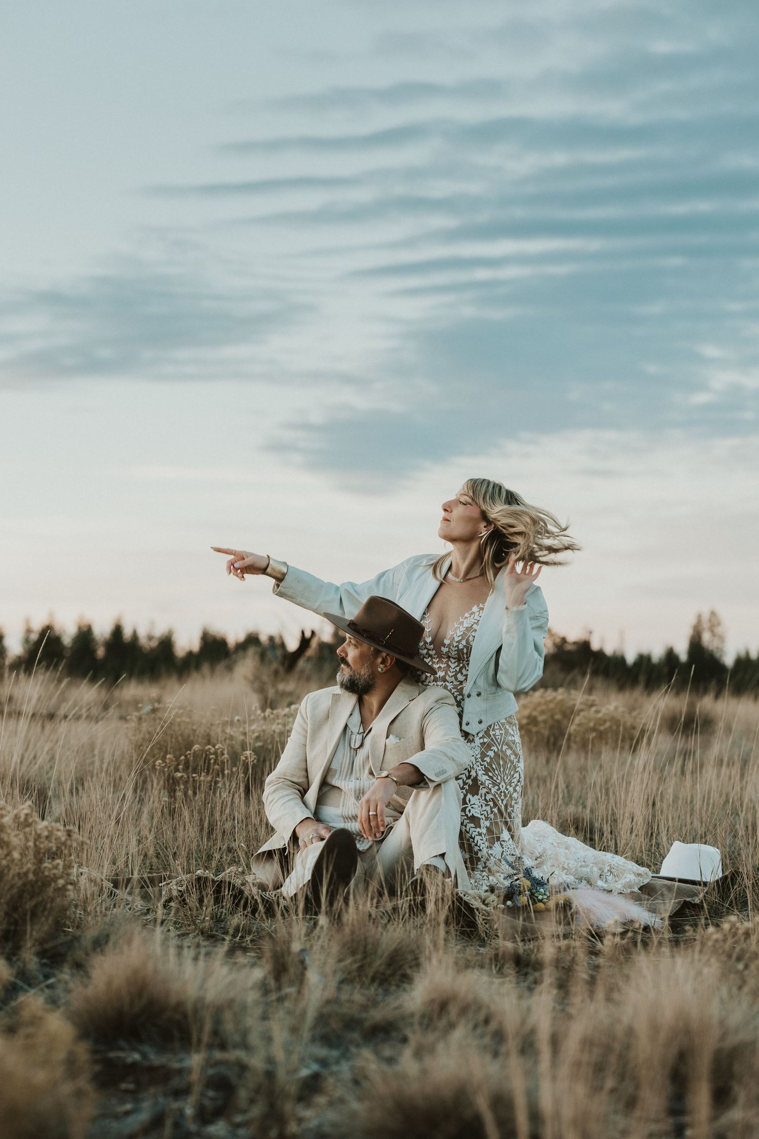 Boho couple sitting in tall grass field at sunset, woman pointing toward the horizon.